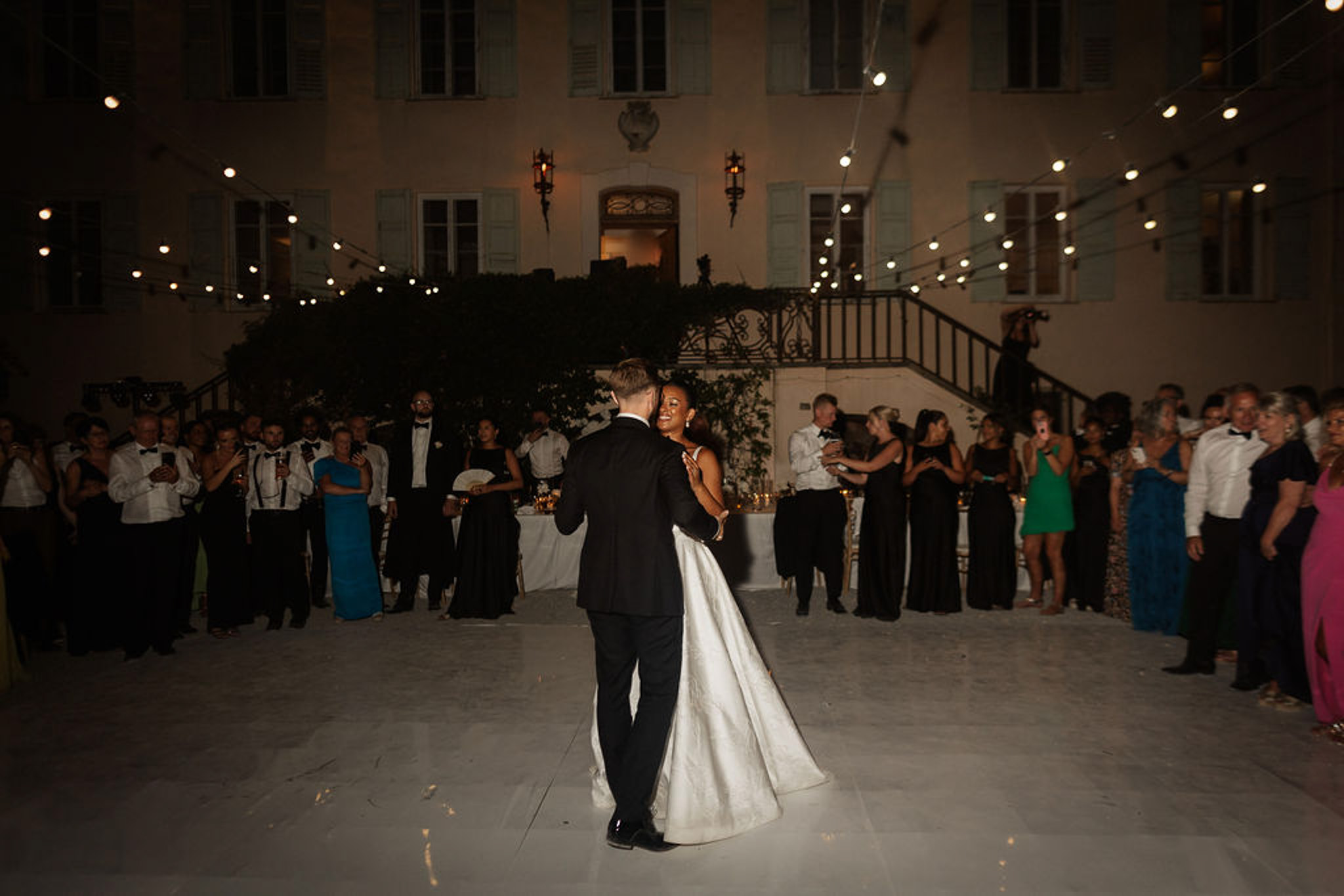 Bride and groom first dance surrounded by guests in elegant neoclassical courtyard at evening reception