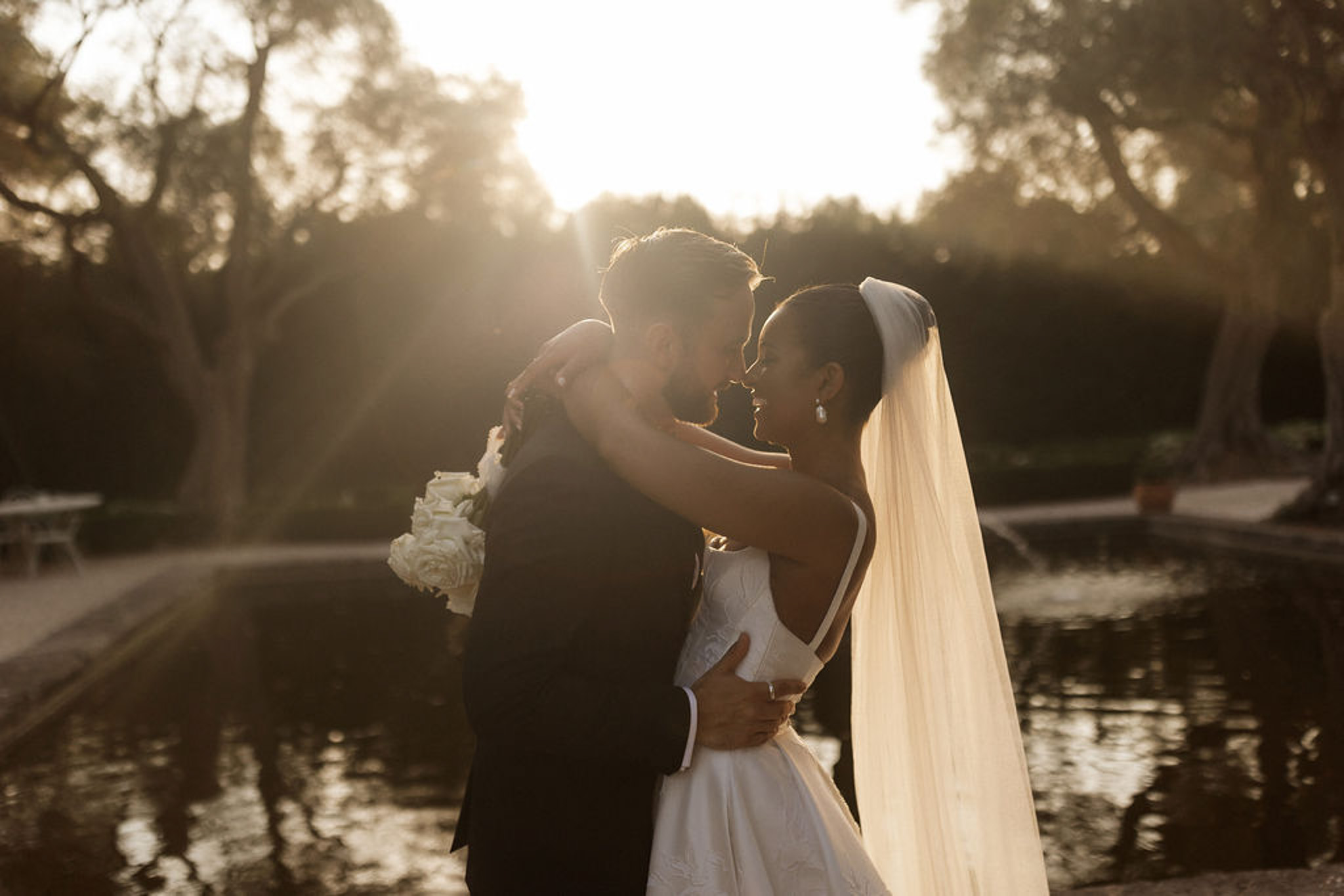 Groom lifting bride during romantic portrait session beside pond in formal garden setting