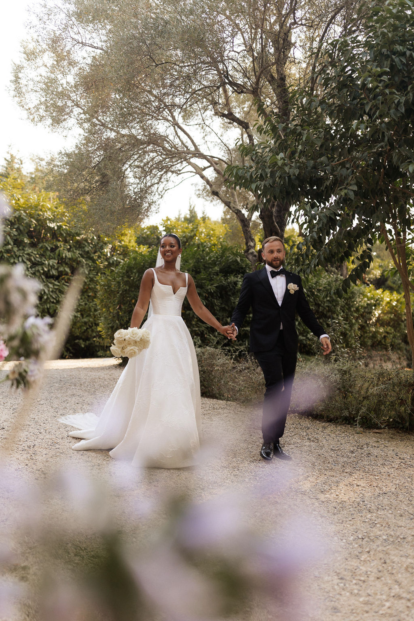 Bride and groom walking hand-in-hand on tree-lined garden pathway during wedding portraits