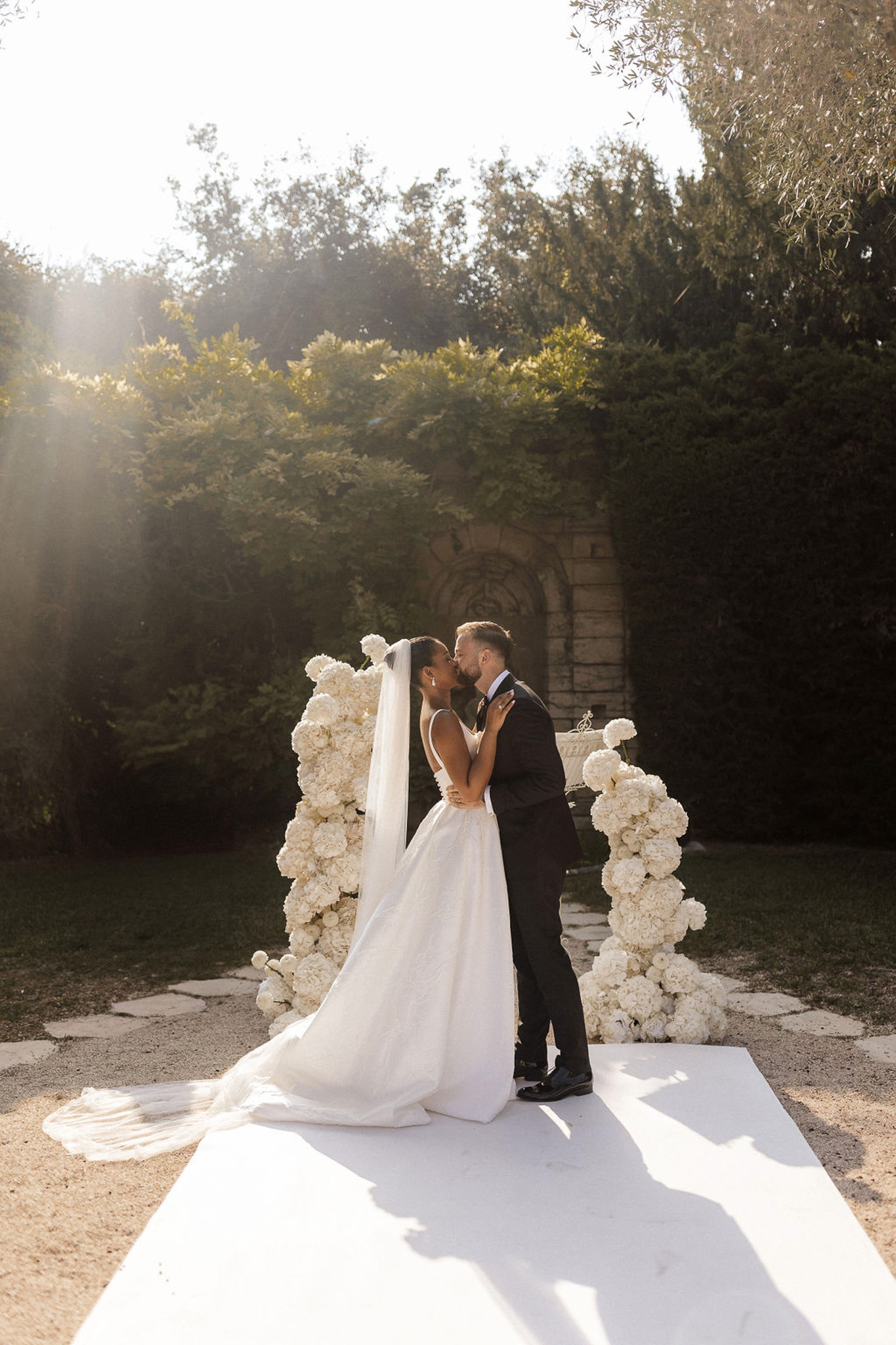 Bride and groom kiss during garden ceremony with white floral archways