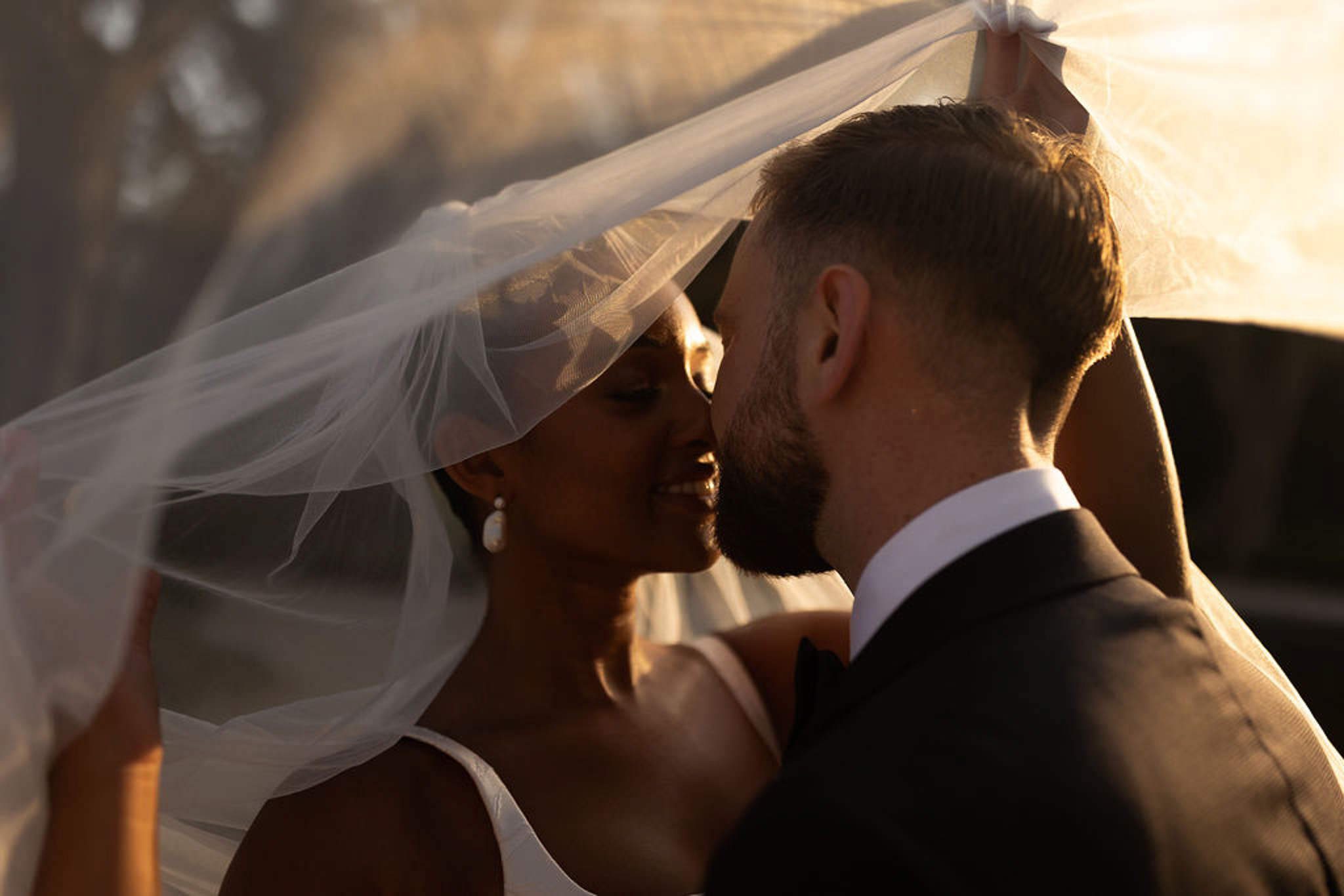 Bride and groom first kiss during outdoor wedding ceremony with ivory veil in golden hour light
