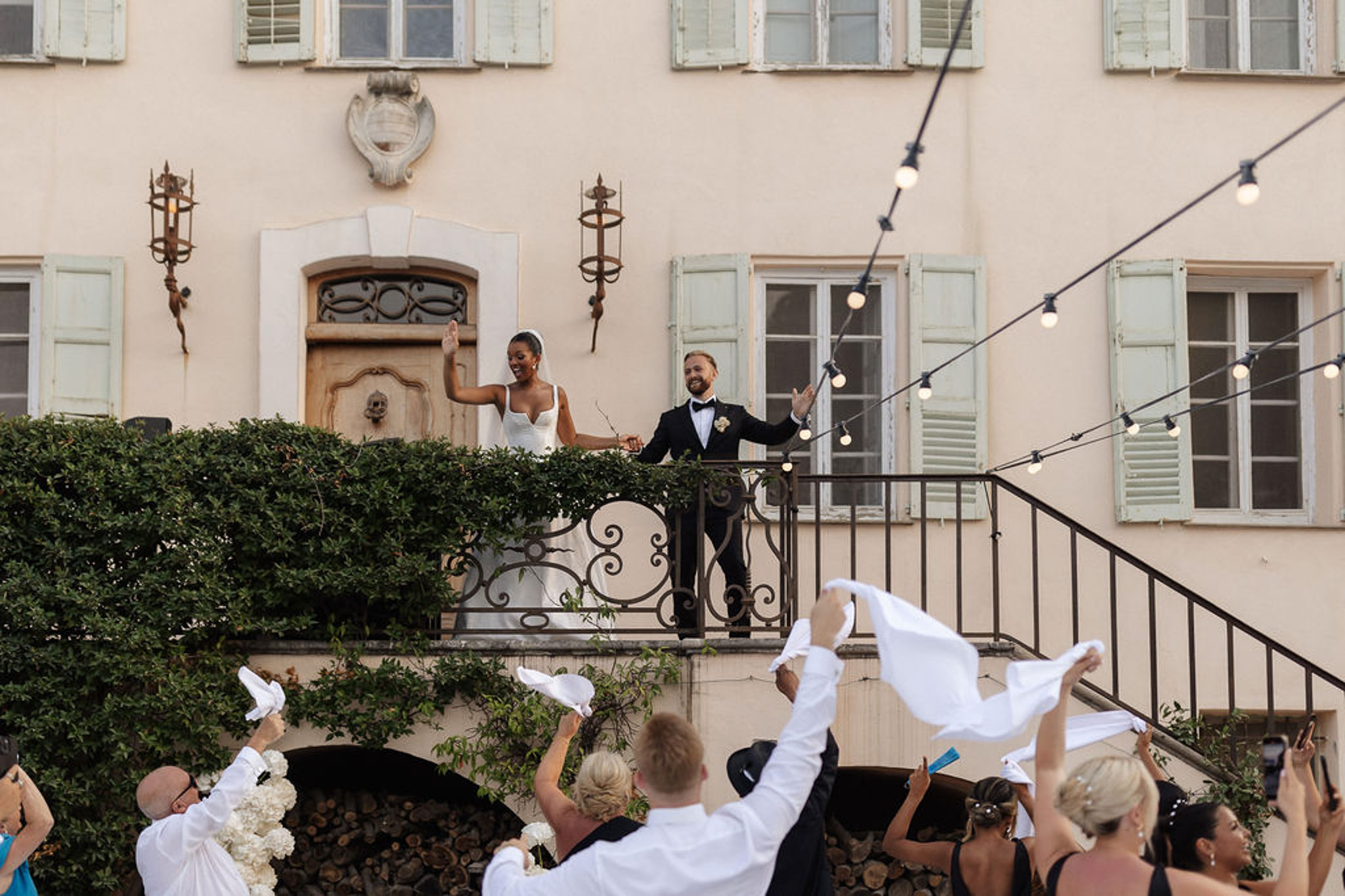 Bride and groom on balcony waving to guests in elegant European-style courtyard with ivy-covered fireplace