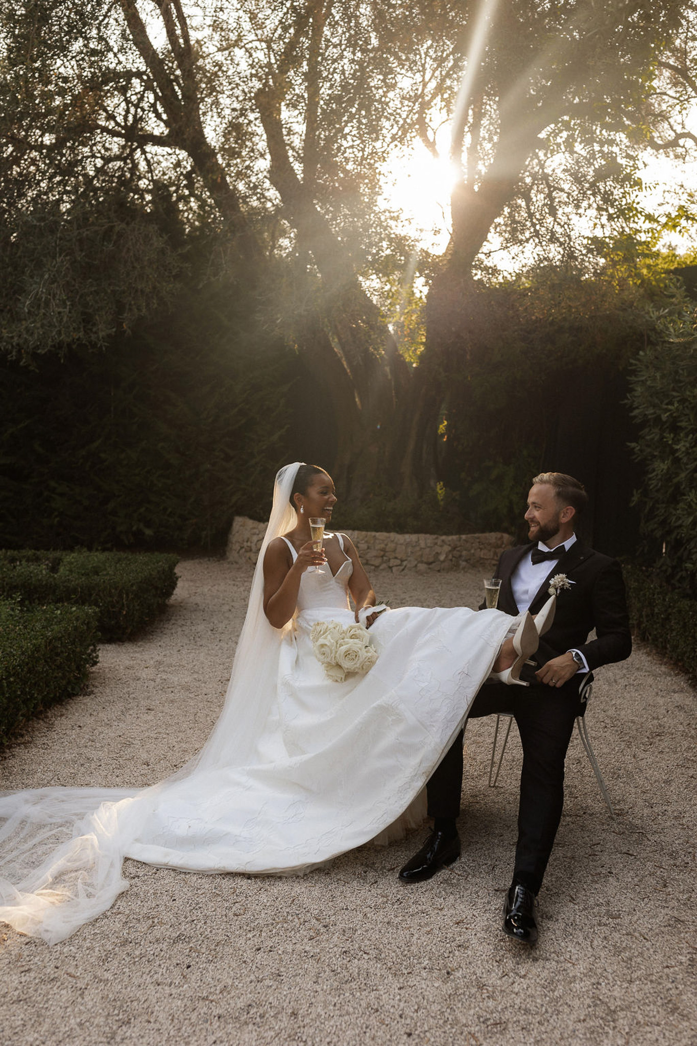 Bride and groom with champagne flutes in tree-lined garden courtyard during golden hour