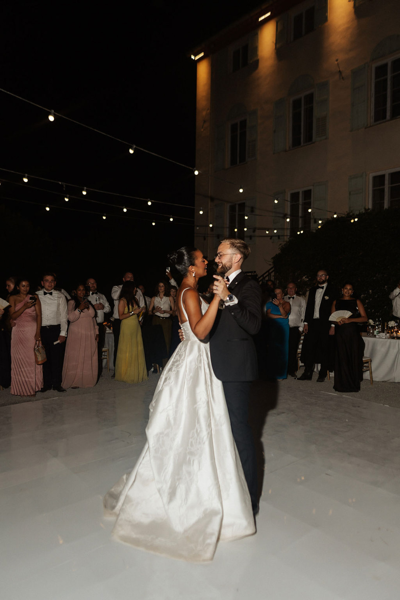 First dance of bride and groom in historic stone courtyard with string lights and guests watching