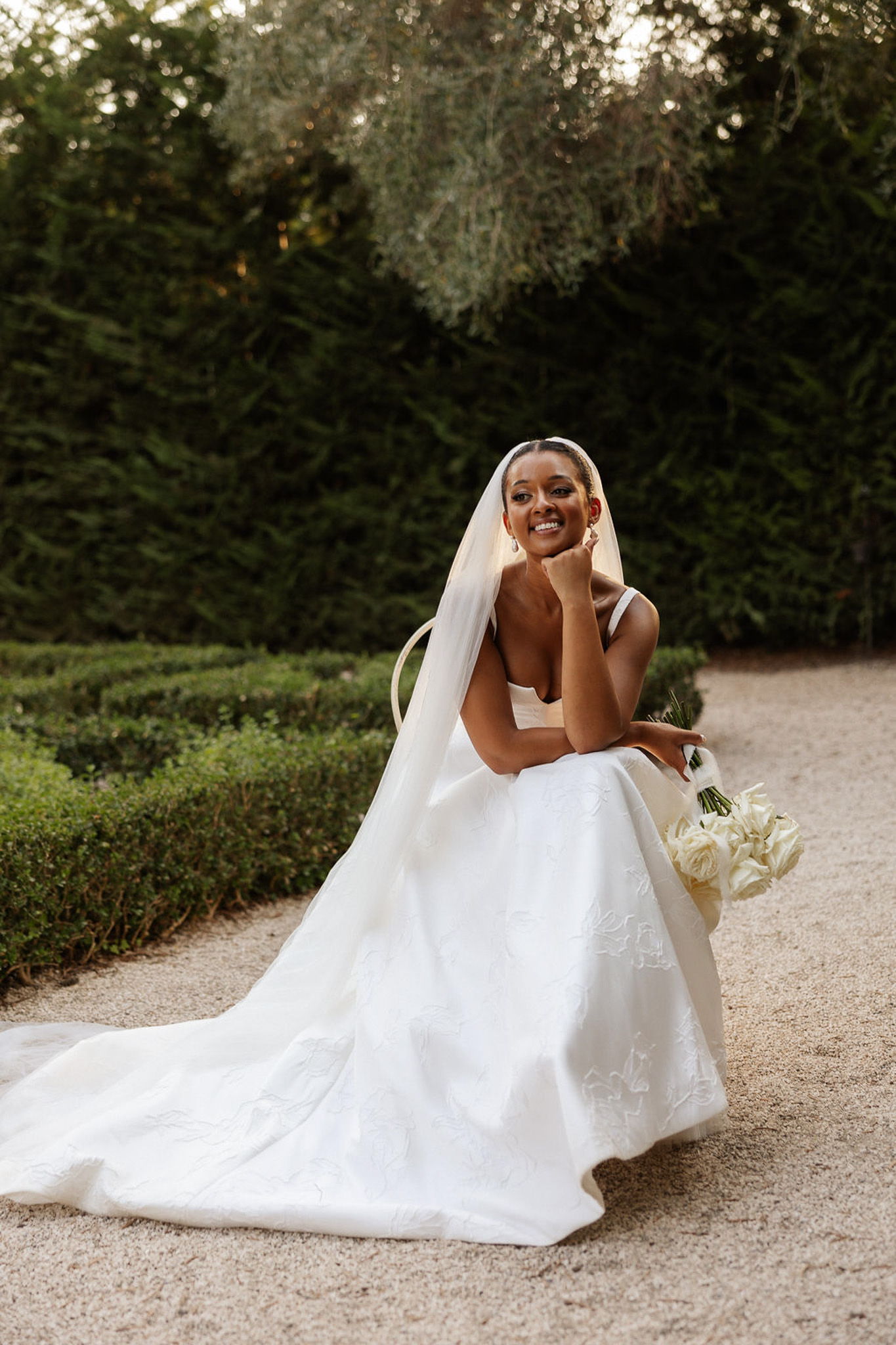 Bride in ivory wedding dress poses in formal garden pathway with manicured hedges and evergreen trees