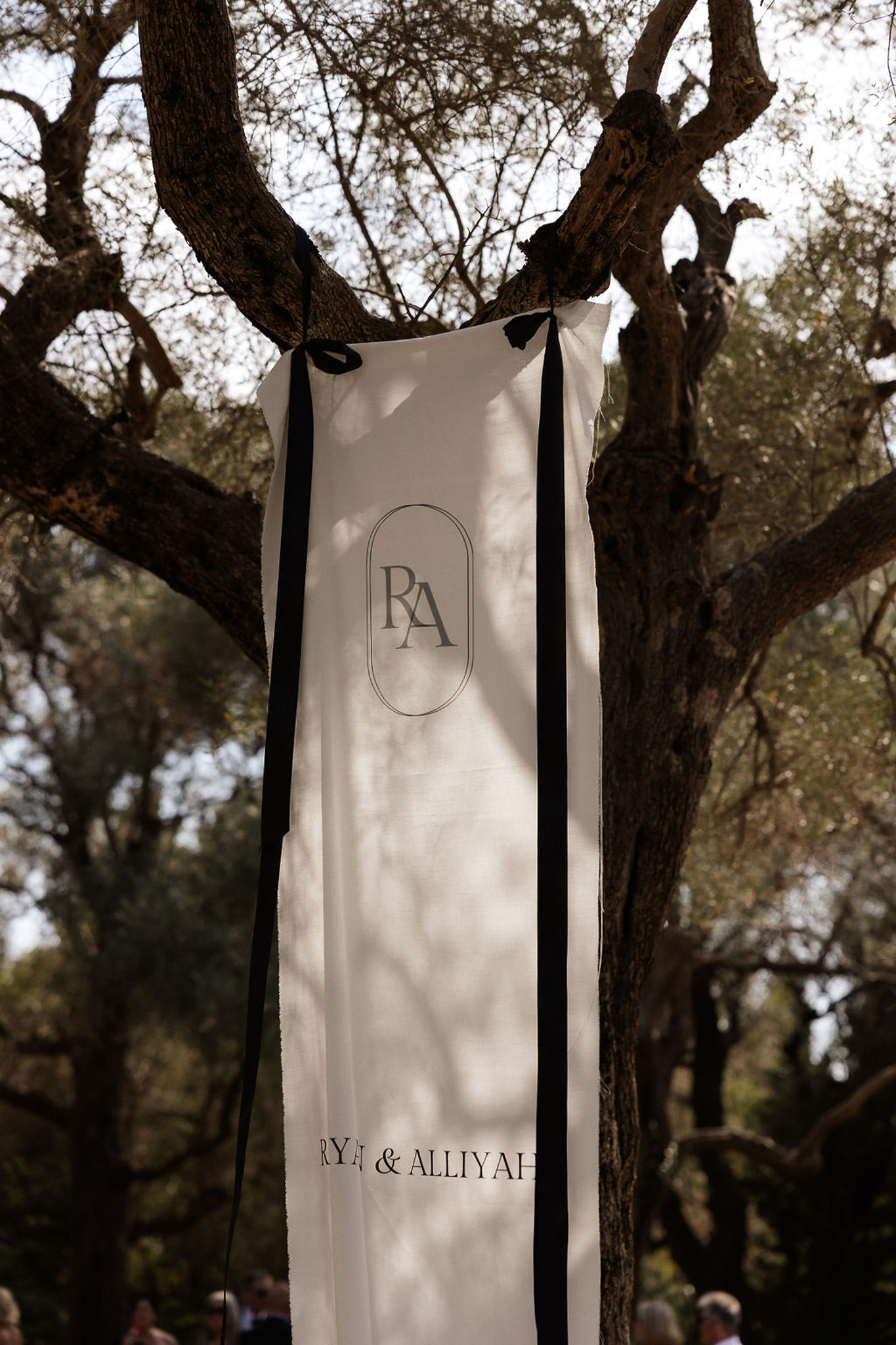 Wedding ceremony banner with couple's monogram hanging from olive tree branches at outdoor wedding venue