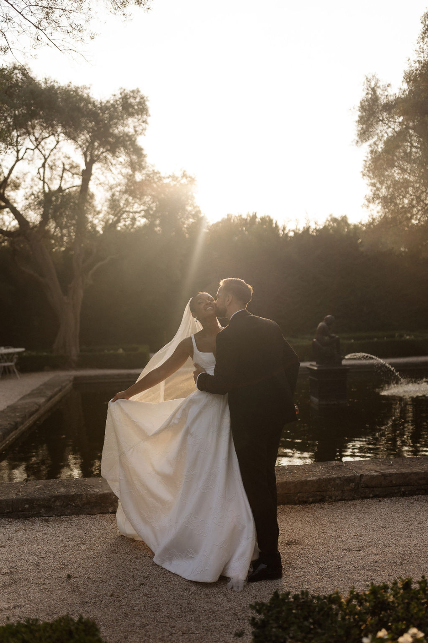 Bride and groom kissing in formal garden during golden hour with water feature and manicured hedges