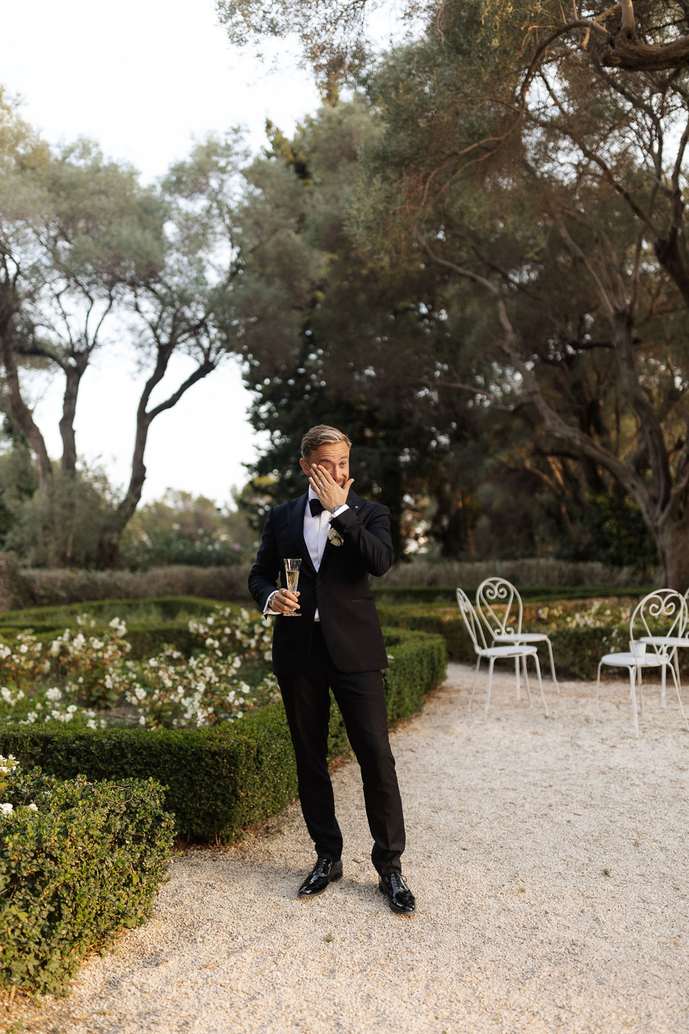 Groom in black tuxedo holding champagne flute during cocktail hour in formal garden with olive trees and boxwood hedges