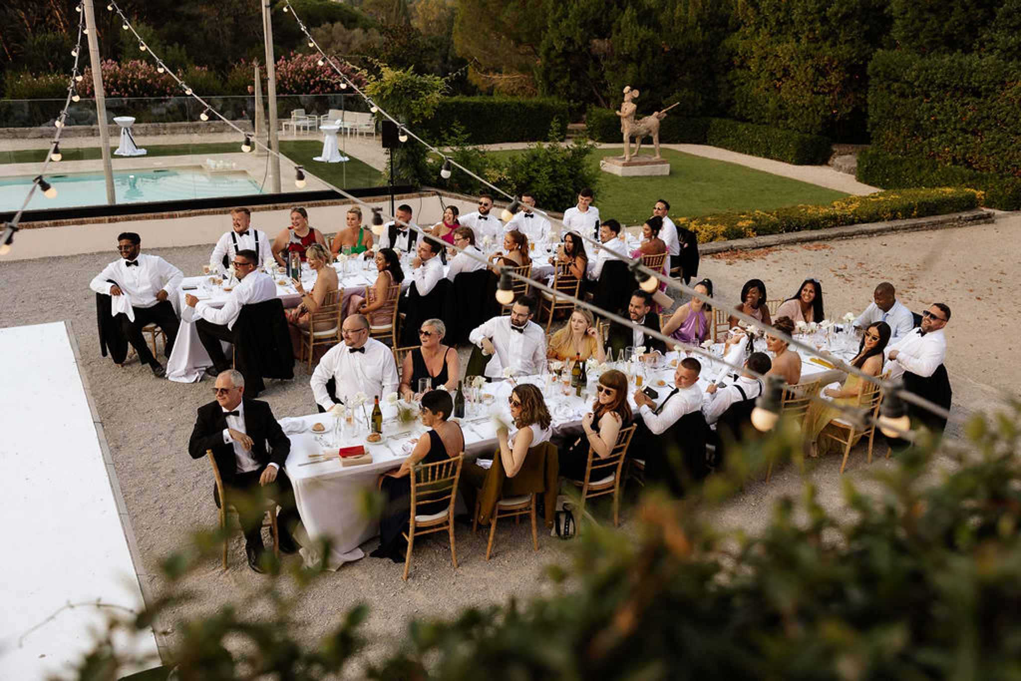 Aerial view of outdoor reception dinner with guests at round tables beside swimming pool at elegant venue