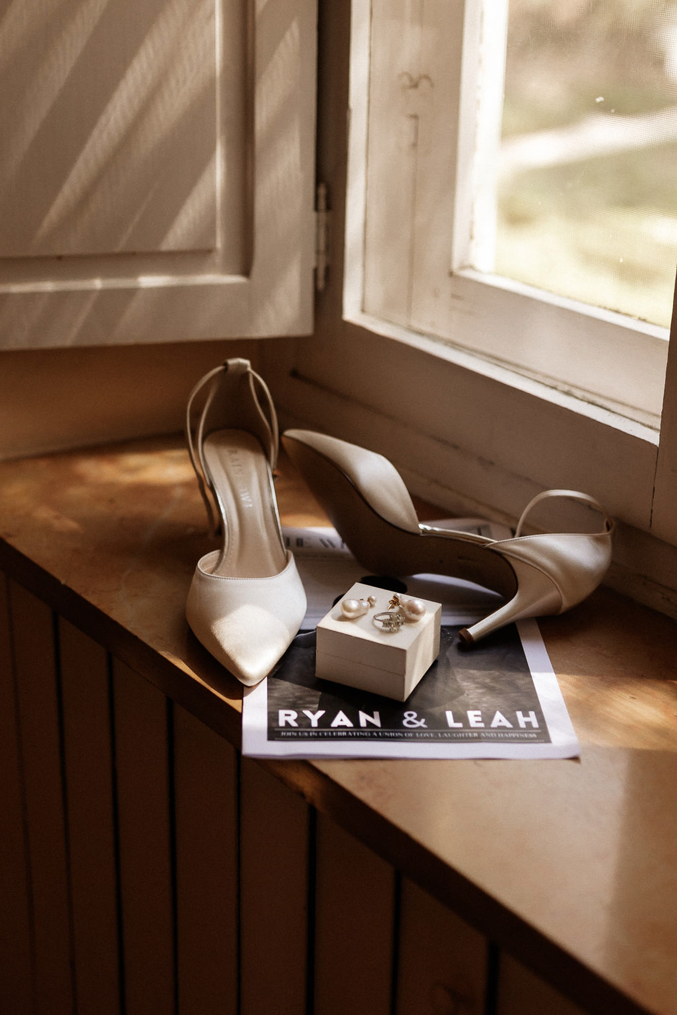 Bridal accessories flat lay with white heels, gold rings, and wedding stationery on wooden windowsill