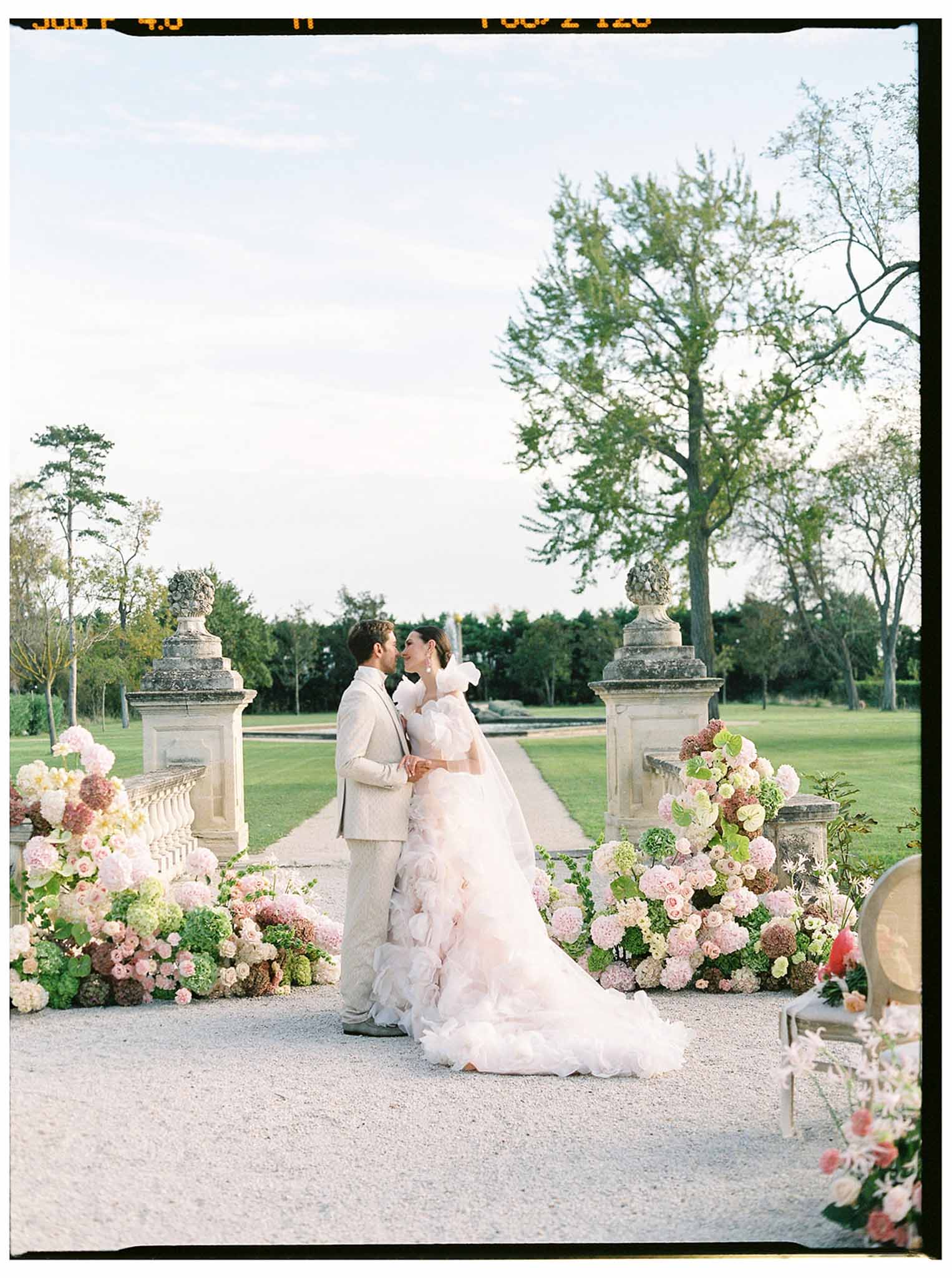 Bride and groom kissing on garden pathway at formal estate with floral arrangements