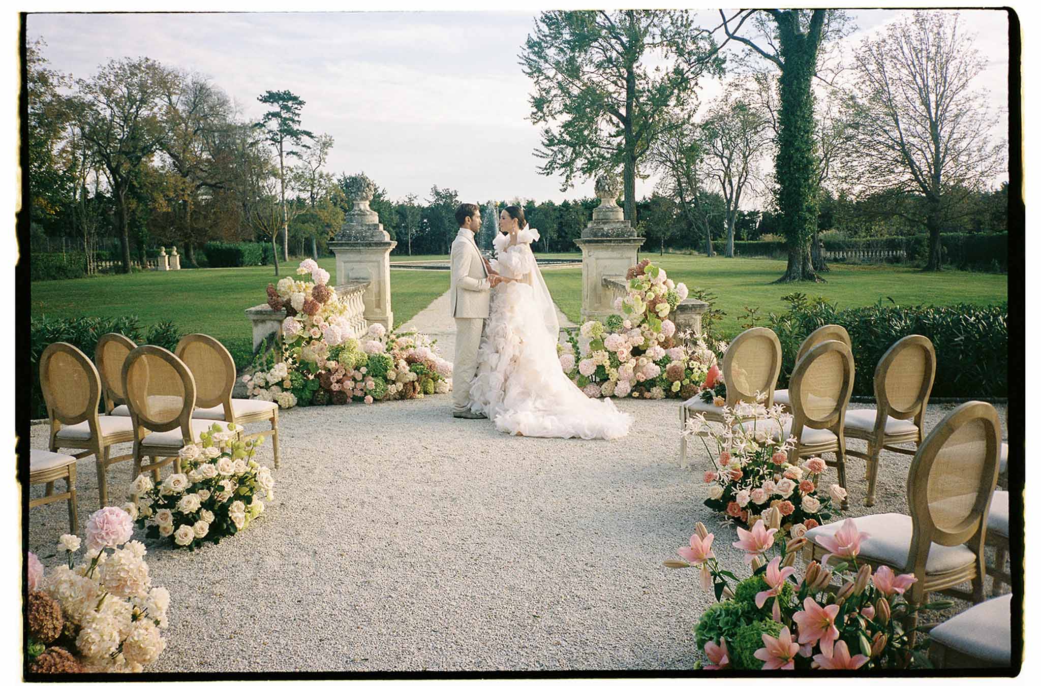 Bride and groom exchanging vows in formal garden ceremony with classical stone columns and manicured lawns