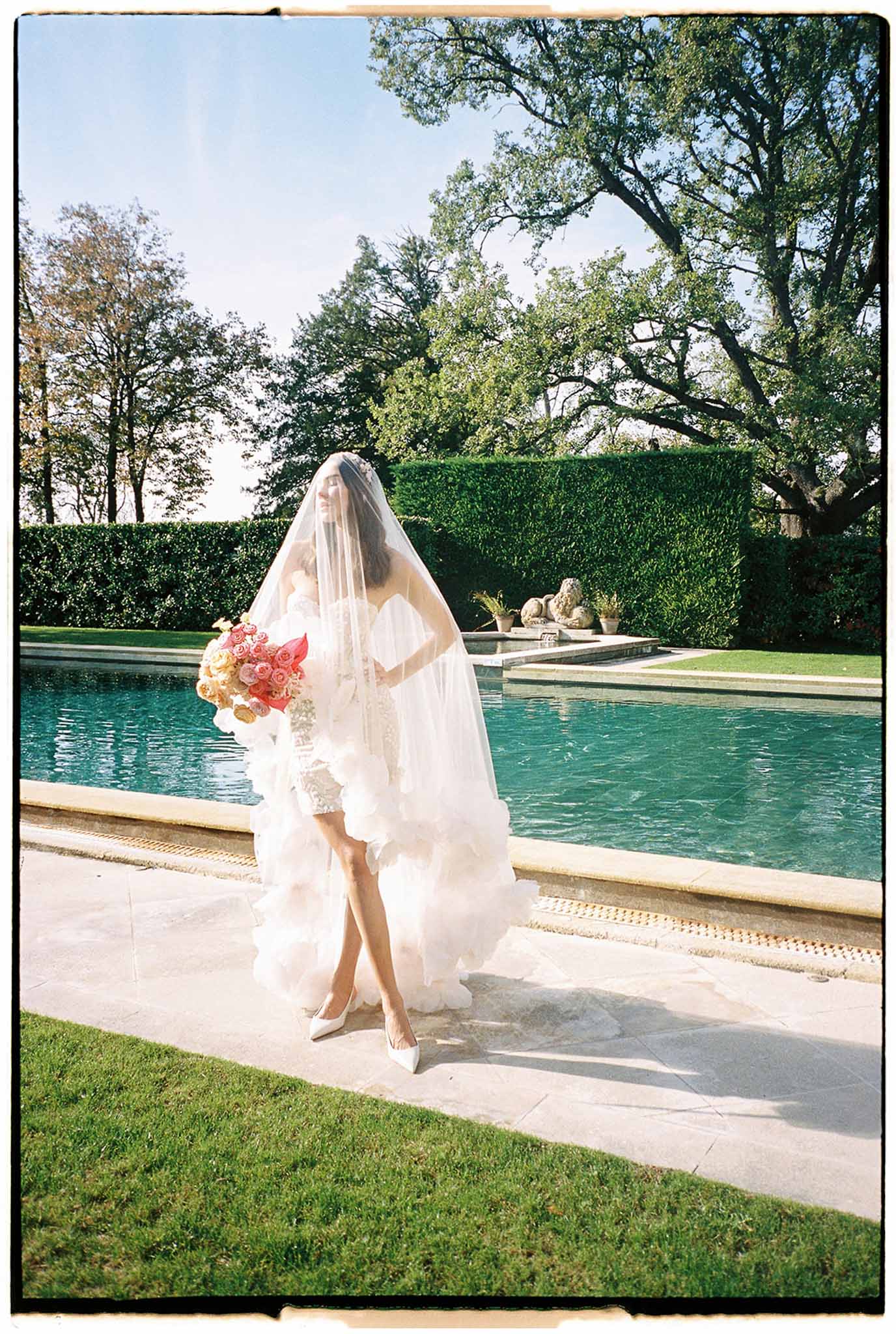 Bride in ivory wedding dress with coral bouquet standing beside pool at garden estate