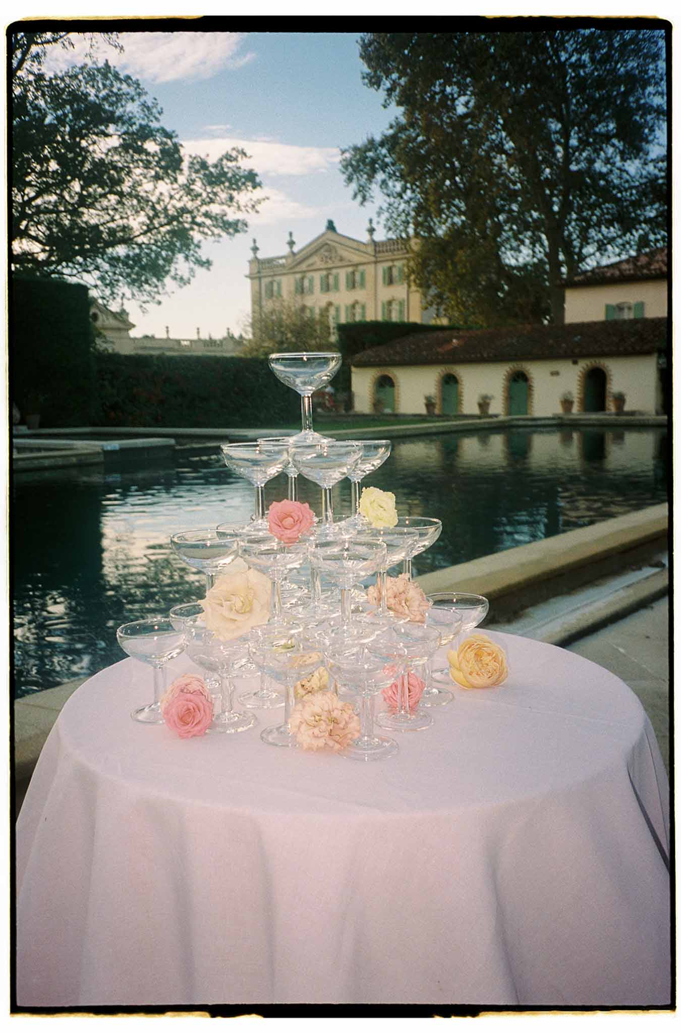 Champagne tower with fresh flowers on lavender linen at waterside terrace overlooking Italian villa