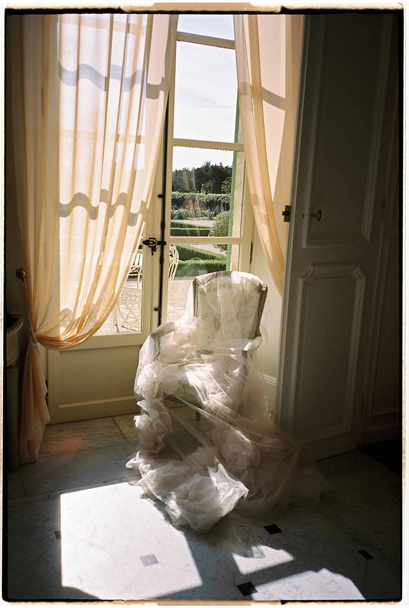Ivory wedding dress draped over chair by French doors with garden view during bridal preparation