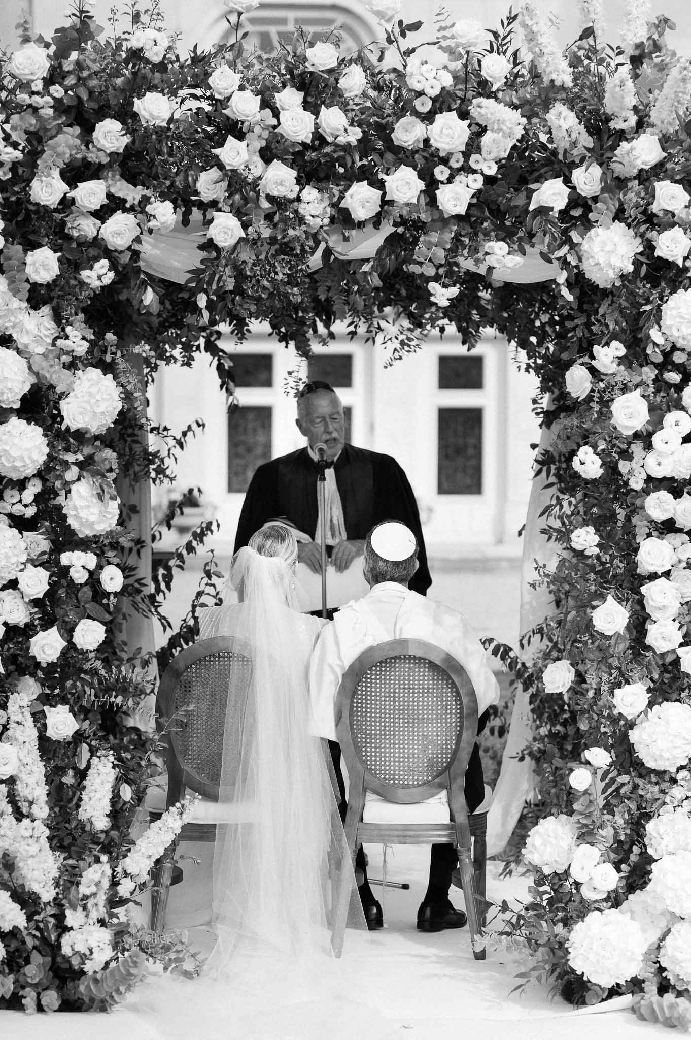 Wedding ceremony under elaborate floral arch at classical venue with bride, groom, and officiant