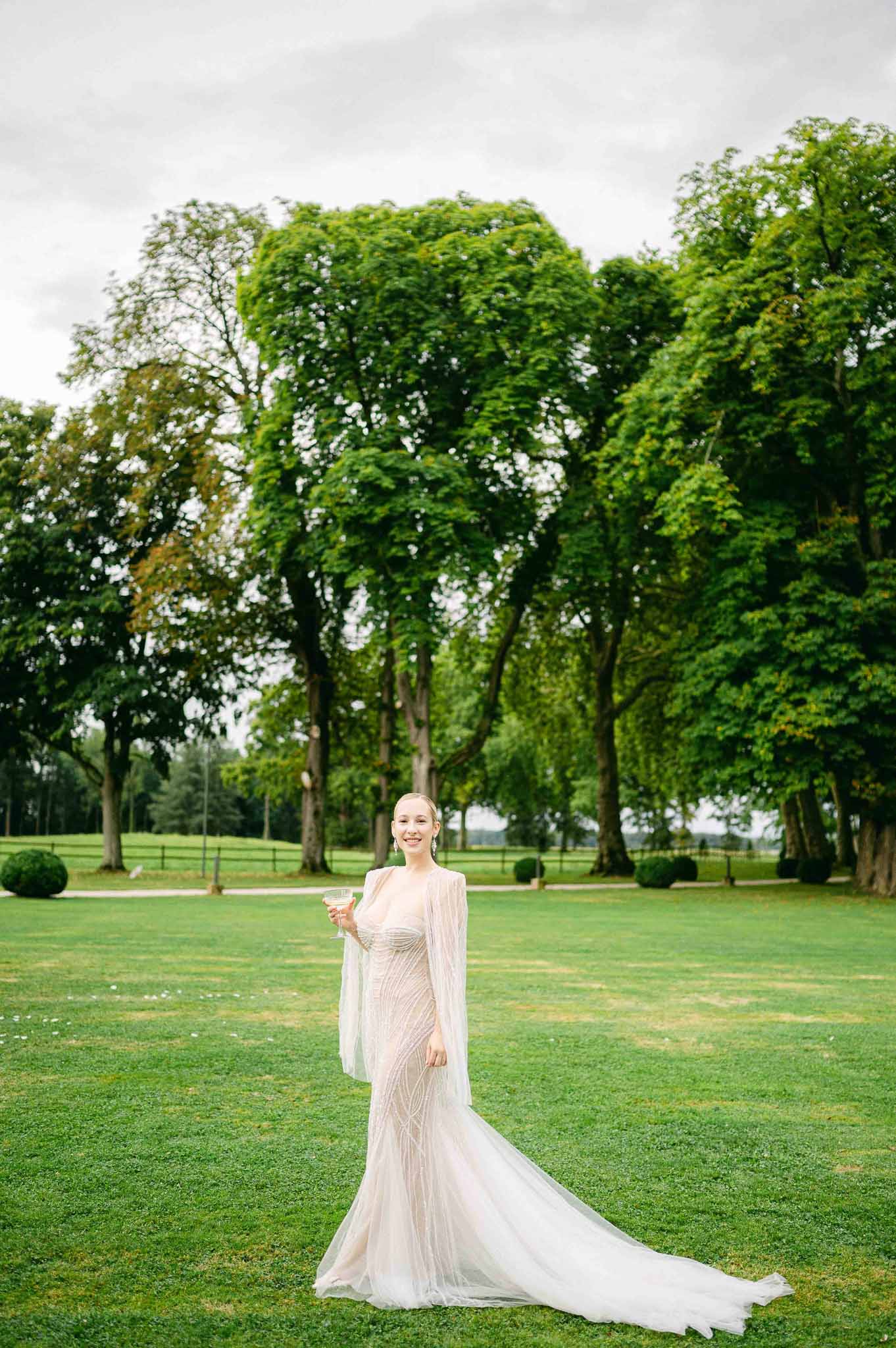 Bride in ivory gown holding champagne flute in manicured estate garden with tree-lined avenue