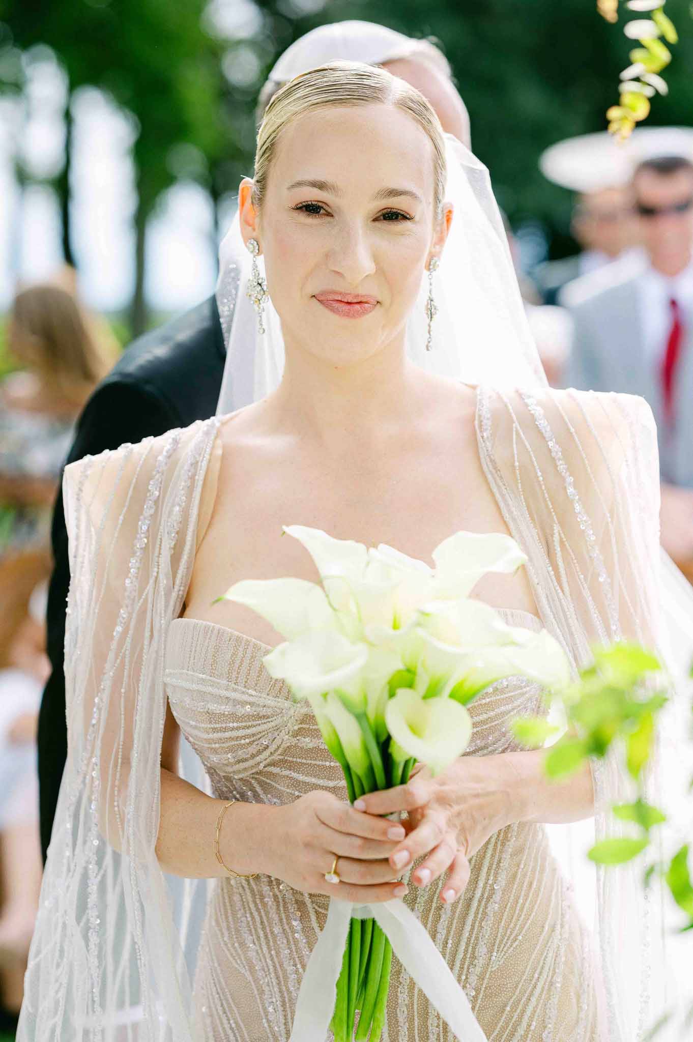 Bride during outdoor garden ceremony holding white calla lily bouquet with guests in background
