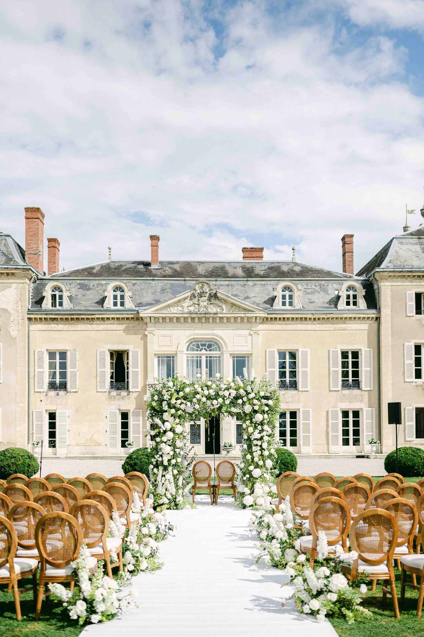 Wedding ceremony setup in French château courtyard with floral arch and wooden chairs