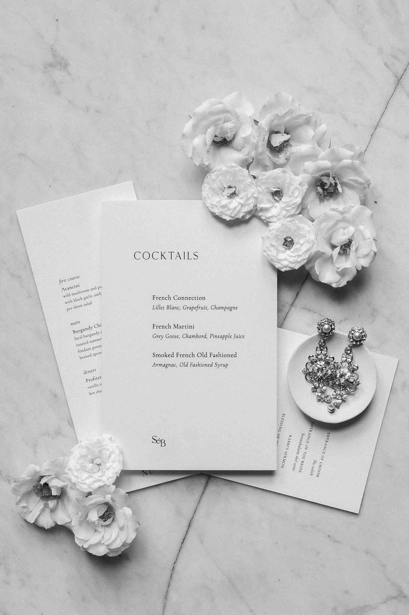 Black and white flat lay of wedding stationery, cocktail menu, white roses and earrings on marble surface