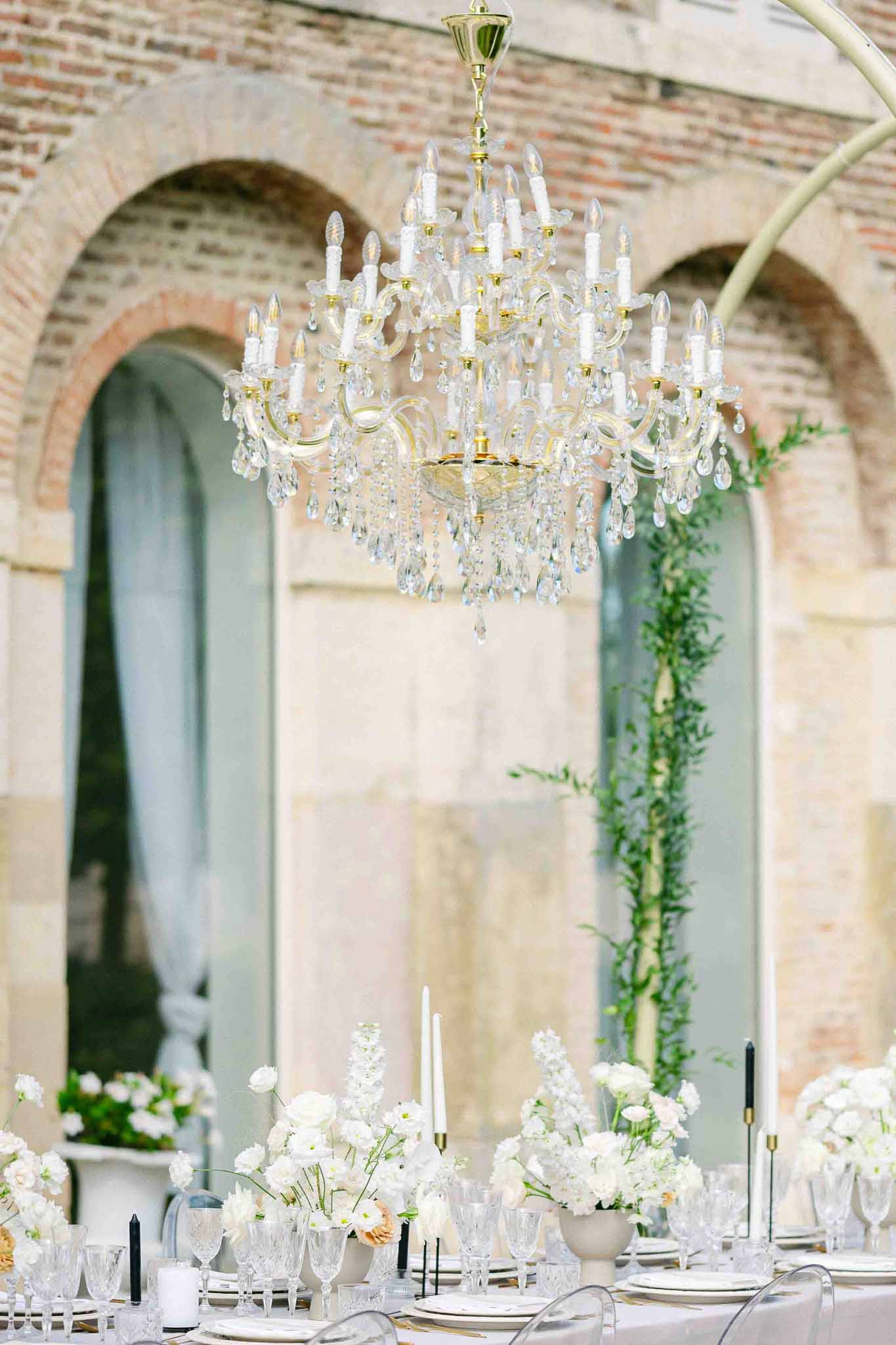 Reception table setup with crystal chandelier and white florals in indoor courtyard with brick archways