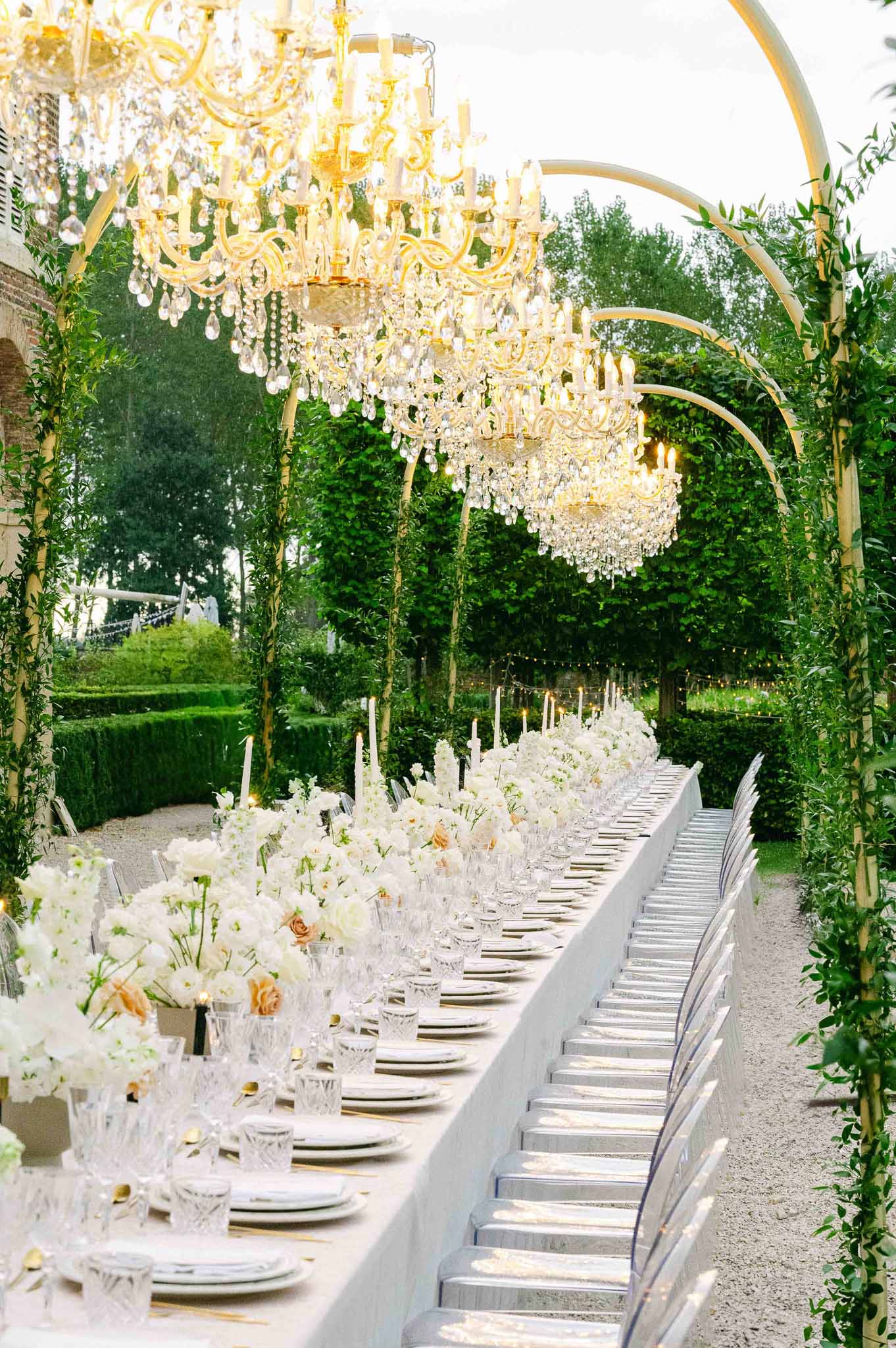 Formal outdoor reception dinner setup with chandeliers in garden courtyard