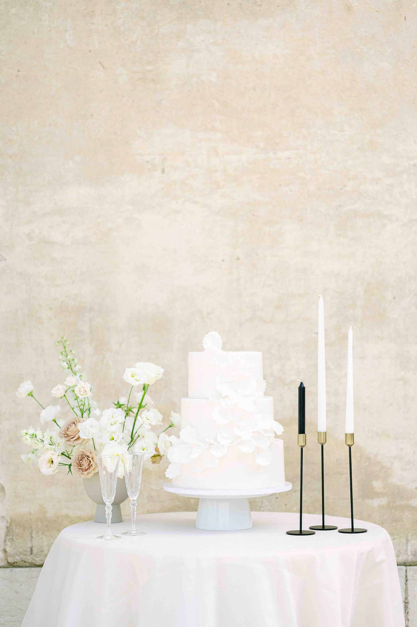 Three-tier white wedding cake with floral arrangement and candlesticks on round table with neutral stone backdrop
