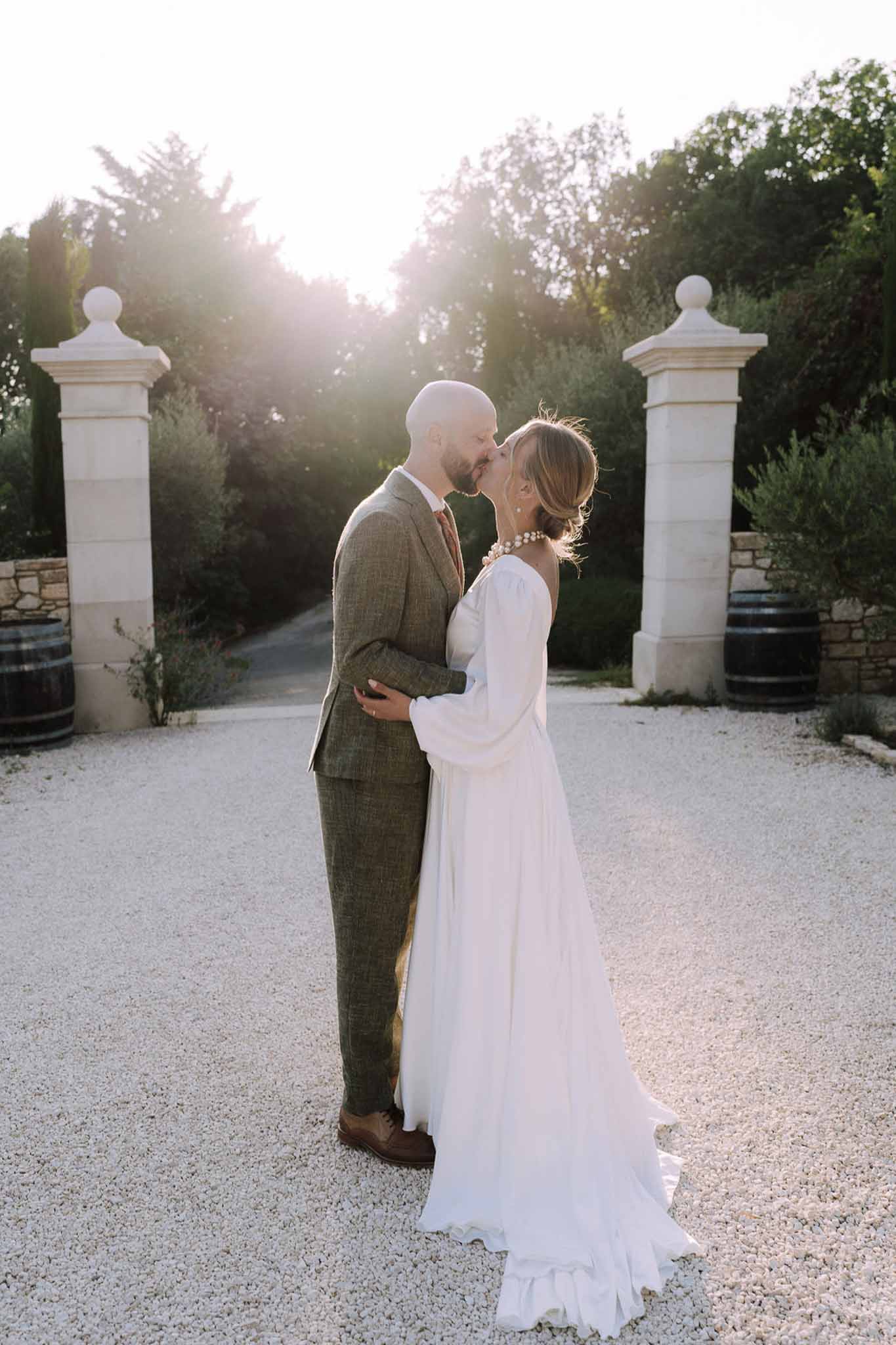 Bride and groom kissing between stone pillars at vineyard estate entrance during golden hour