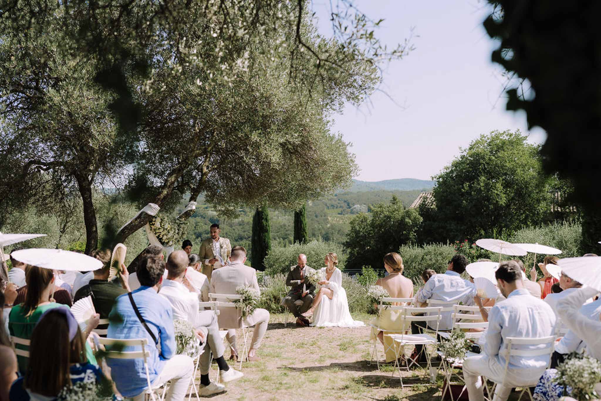 Outdoor wedding ceremony in Tuscan olive grove with guests in wooden chairs and cypress trees