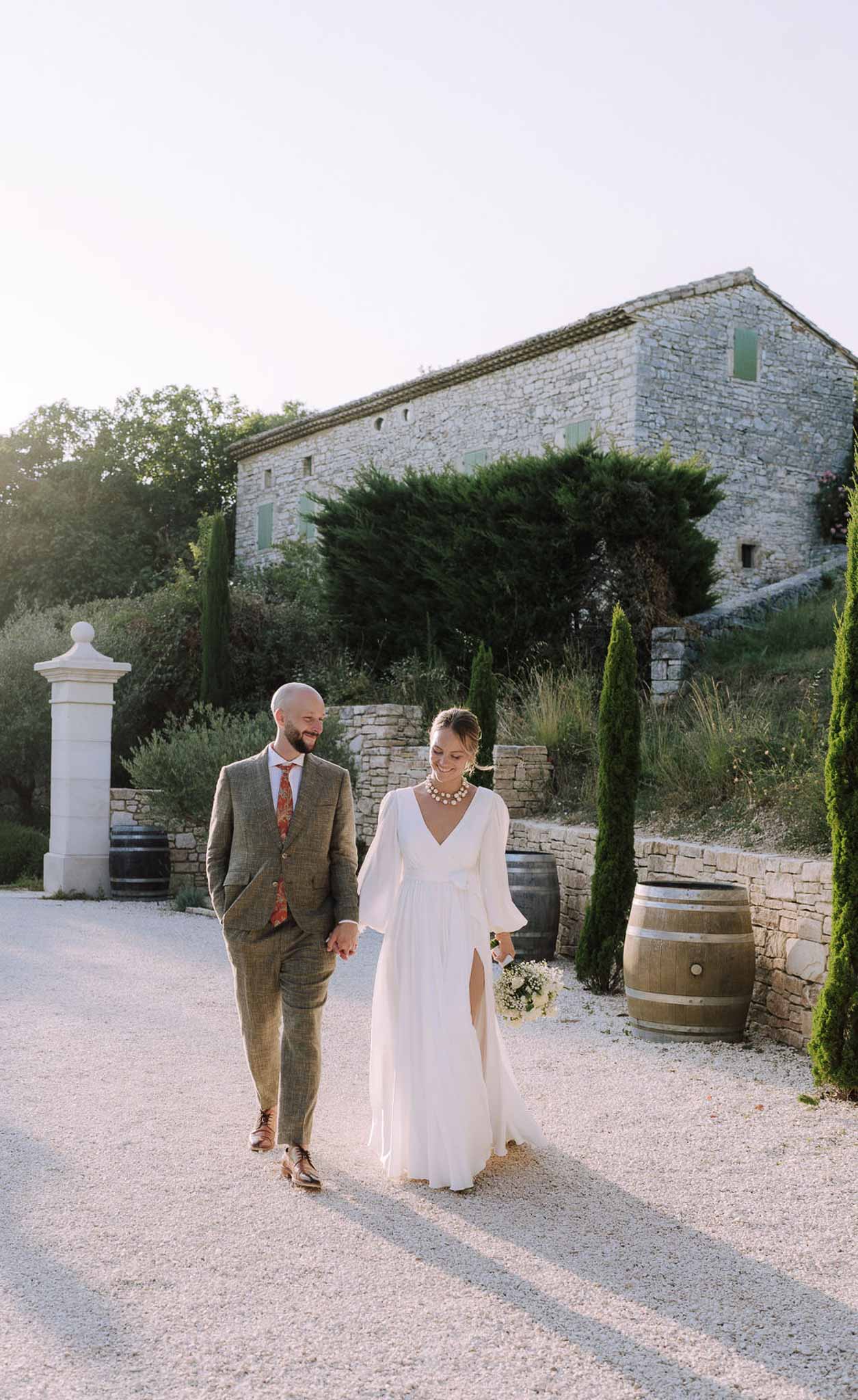 Bride and groom walking hand-in-hand in rustic stone courtyard with cypress trees