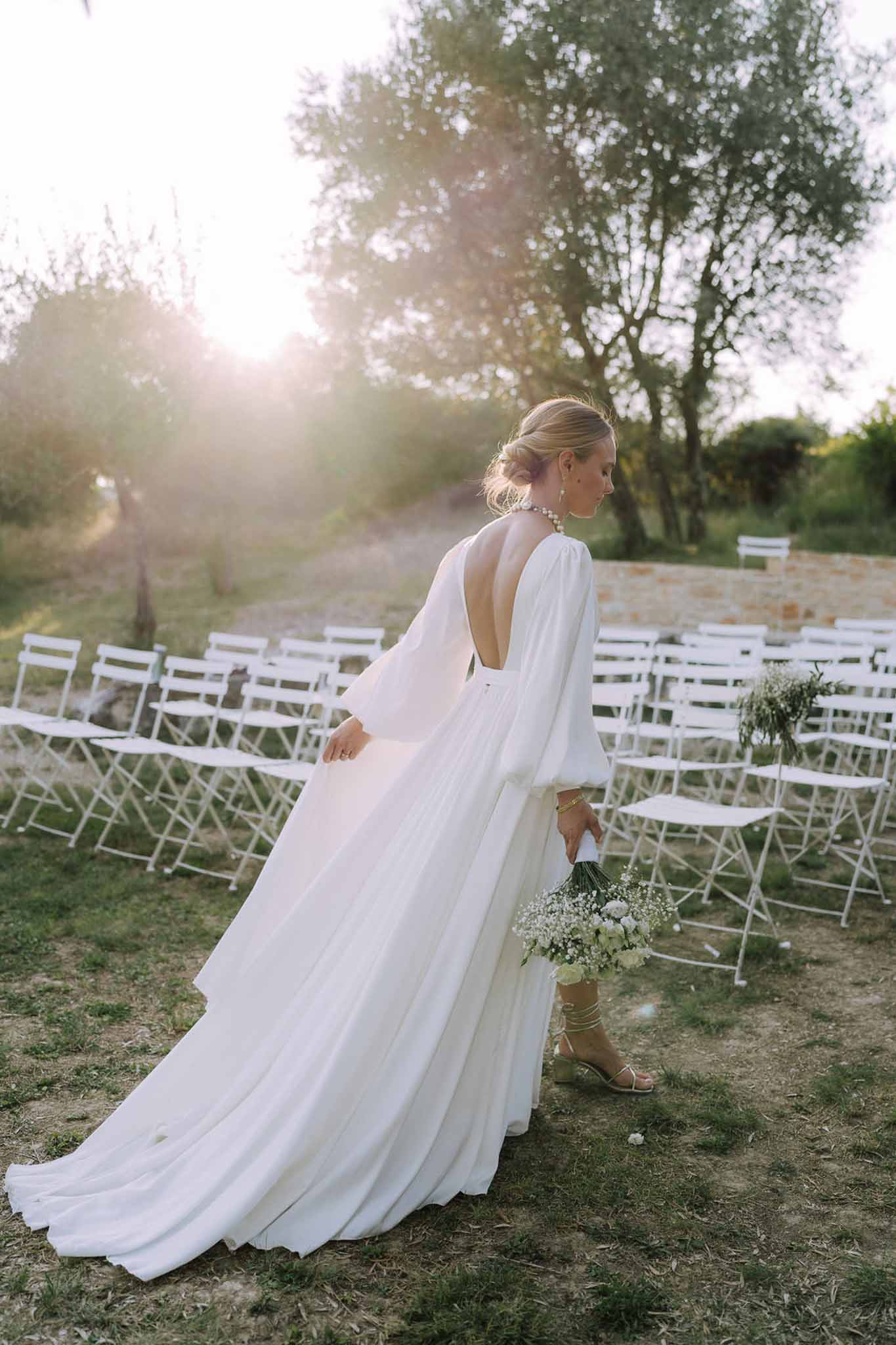 Bride in ivory cape dress holding white bouquet at outdoor ceremony venue with golden light