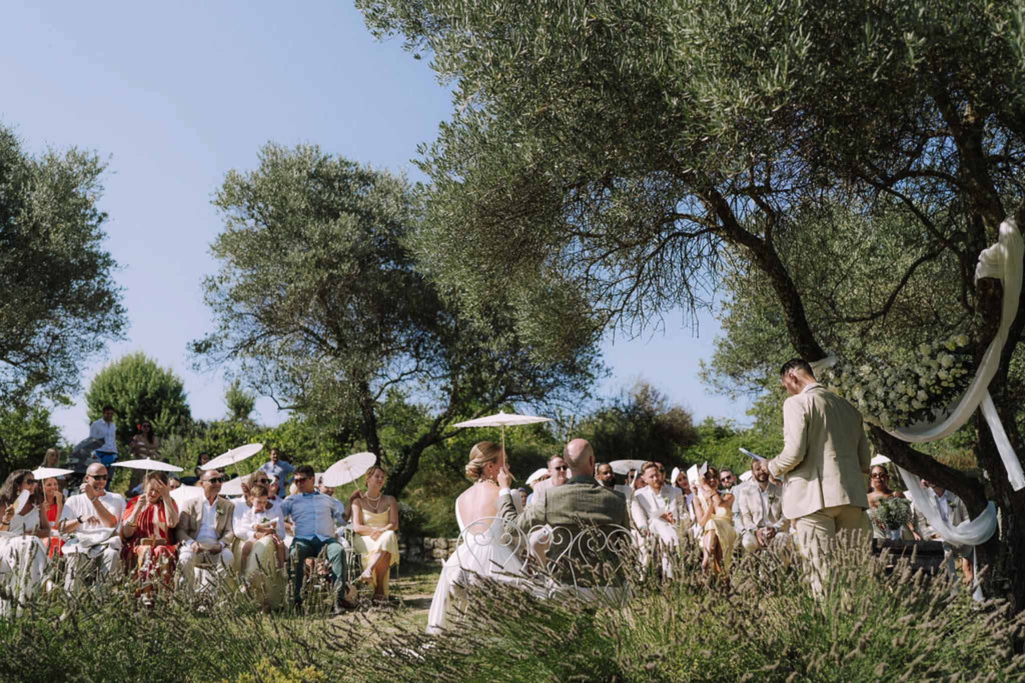 Outdoor wedding ceremony with white arch and guests seated in olive grove garden setting