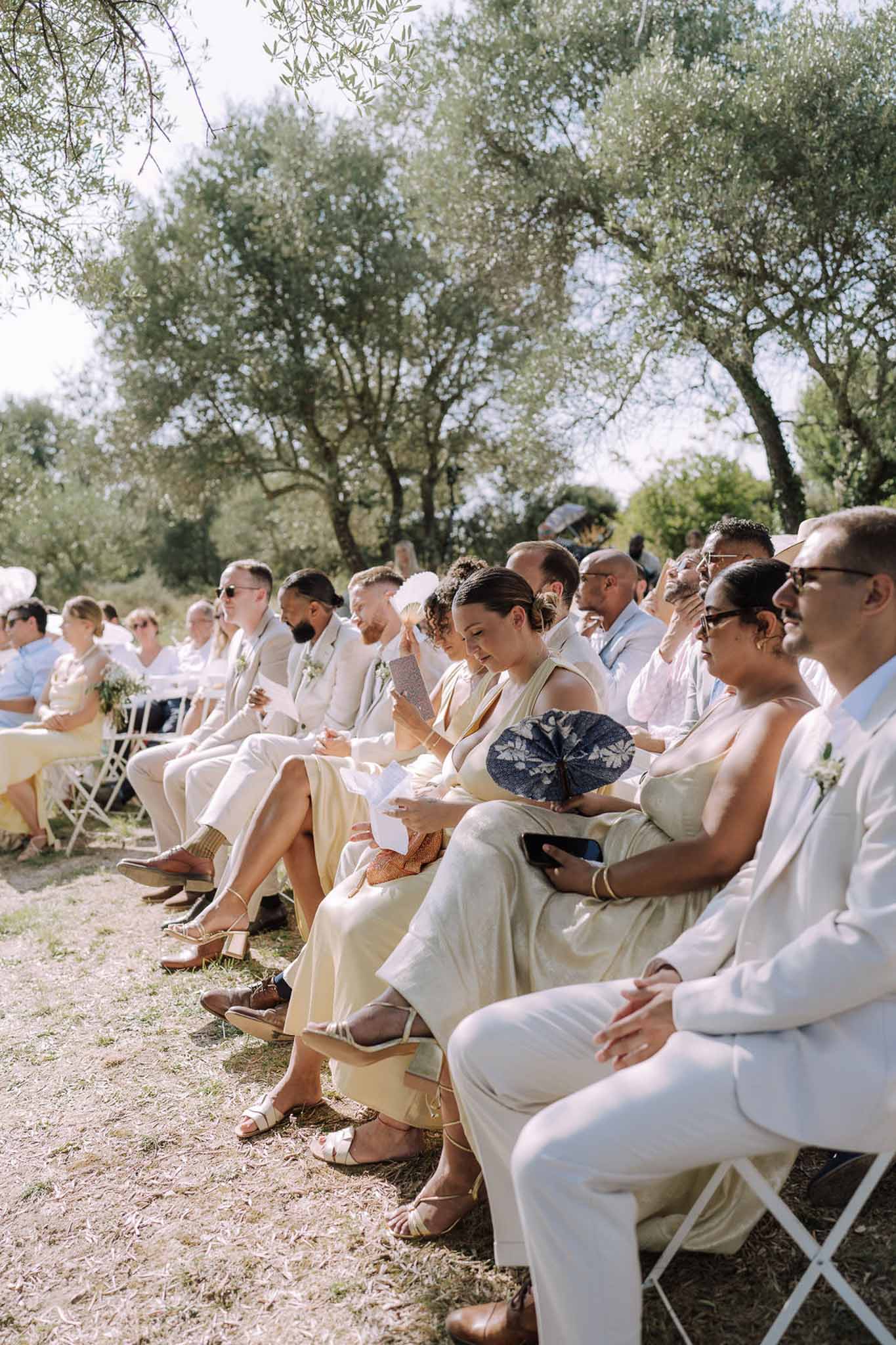 Outdoor wedding ceremony in olive grove with guests in neutral attire seated in white chairs