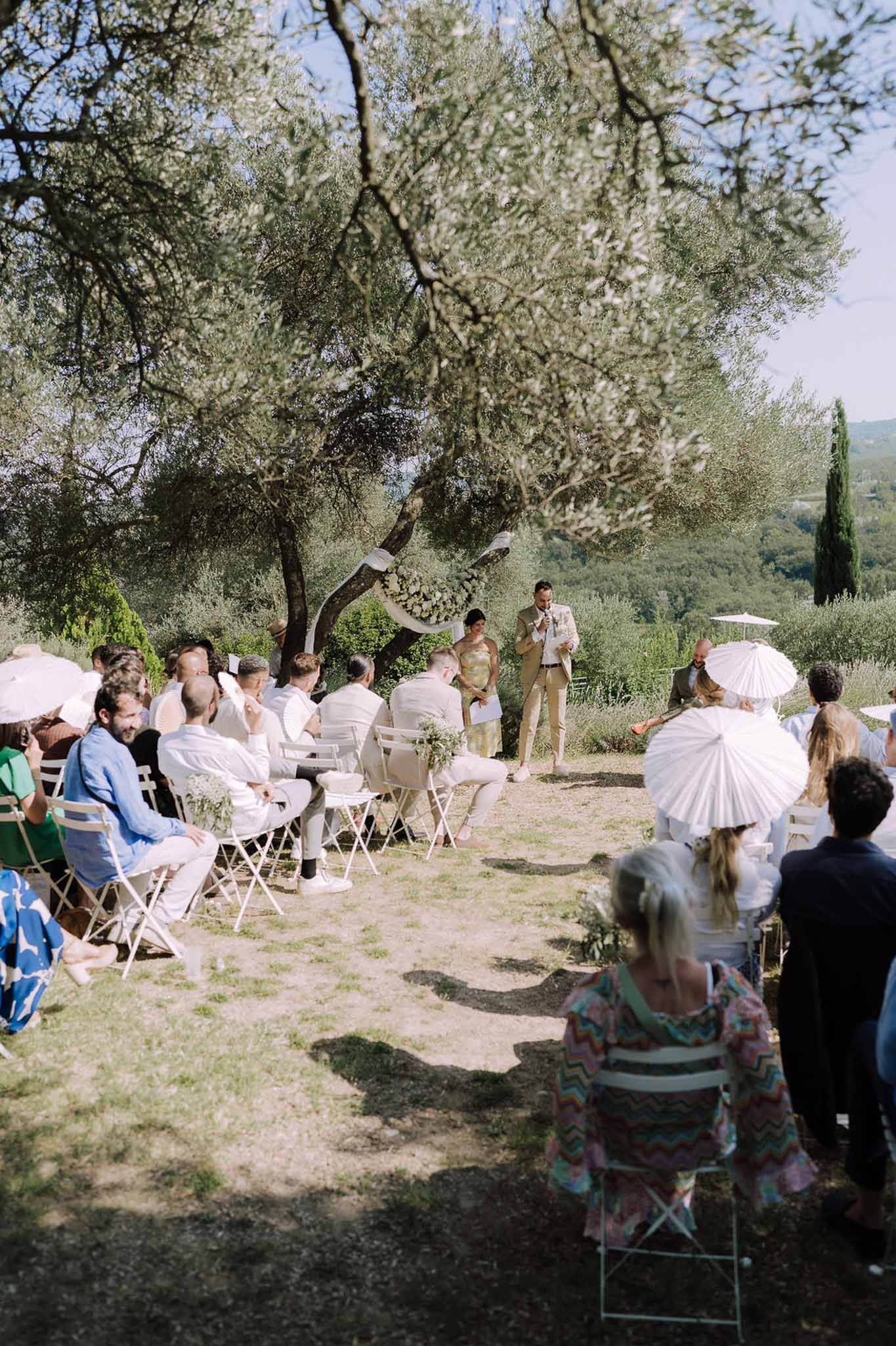 Outdoor wedding ceremony under olive trees with guests holding white umbrellas in Mediterranean setting