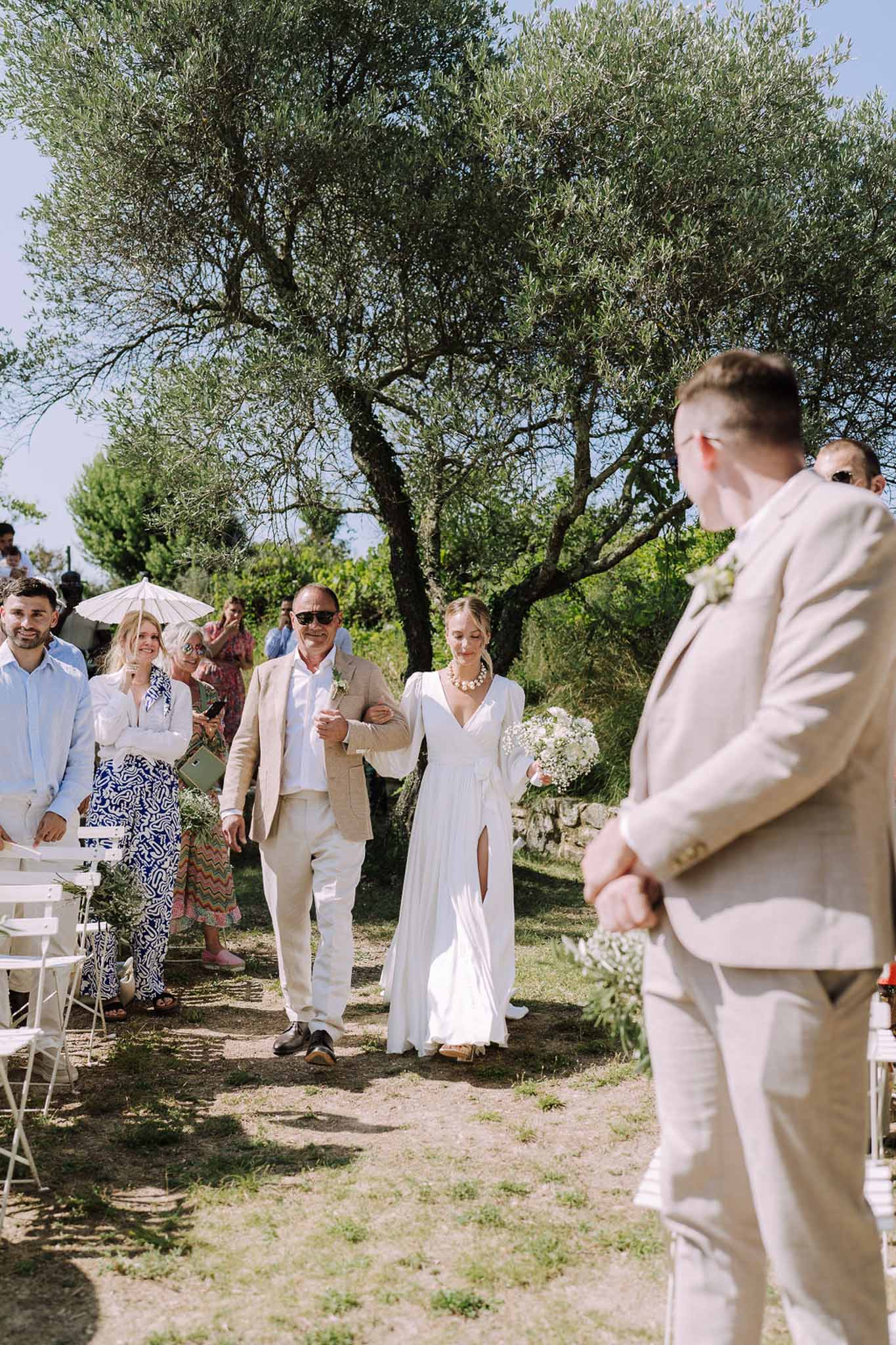 Bride and groom walking down tree-lined aisle during outdoor garden ceremony with guests seated on white chairs