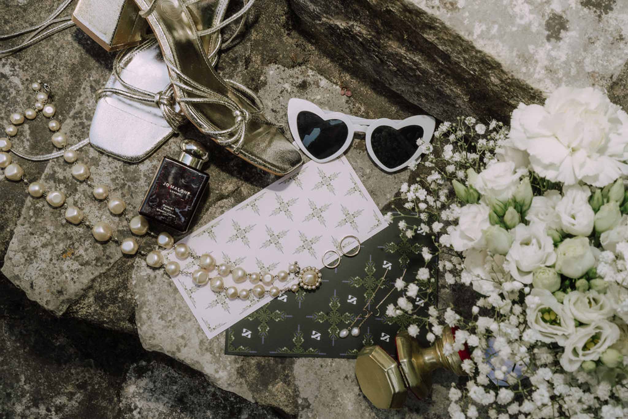 Wedding accessories flat lay with rings, pearls, and white flowers on weathered stone surface