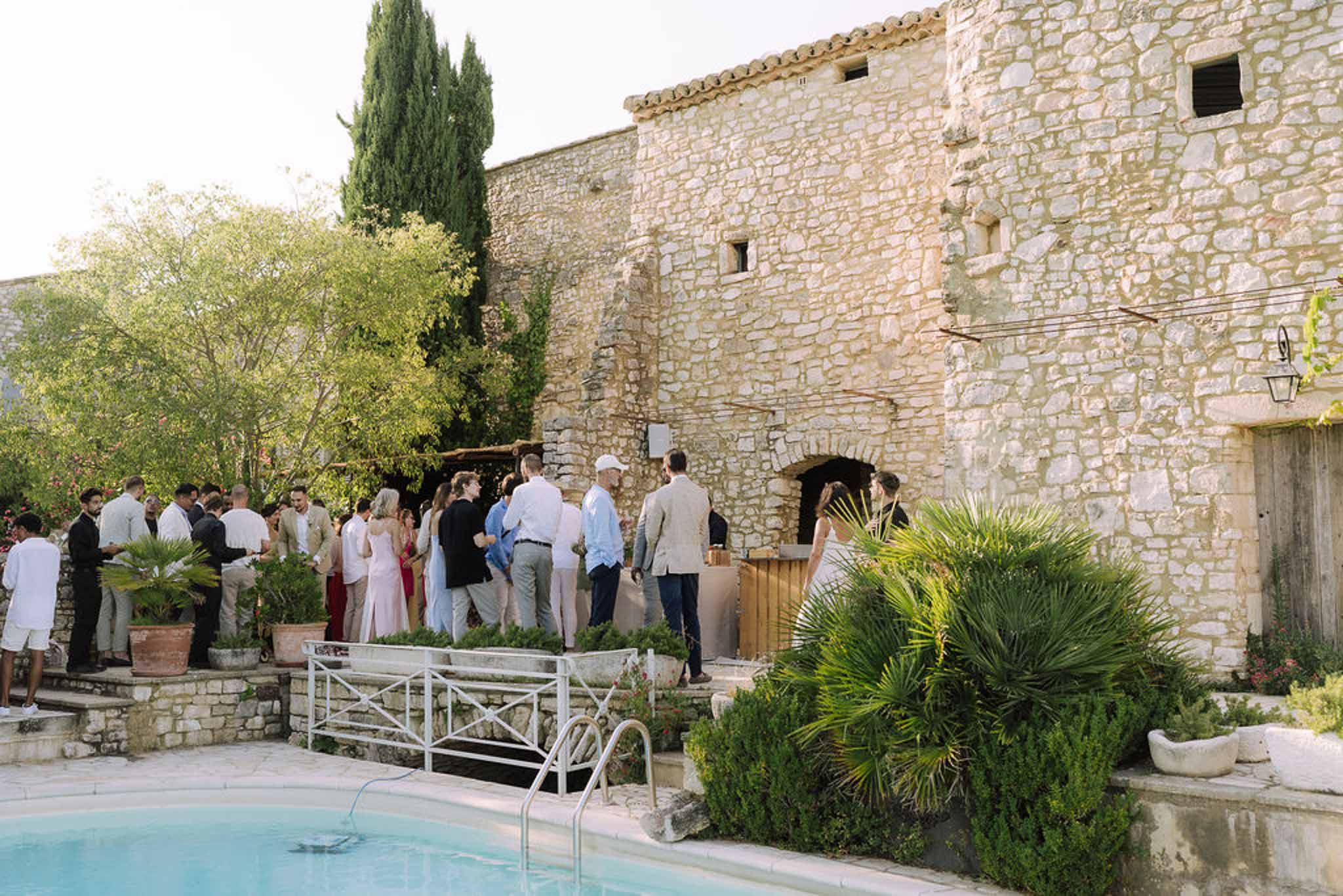 Cocktail hour guests mingling in courtyard of stone villa with pool and Mediterranean landscaping