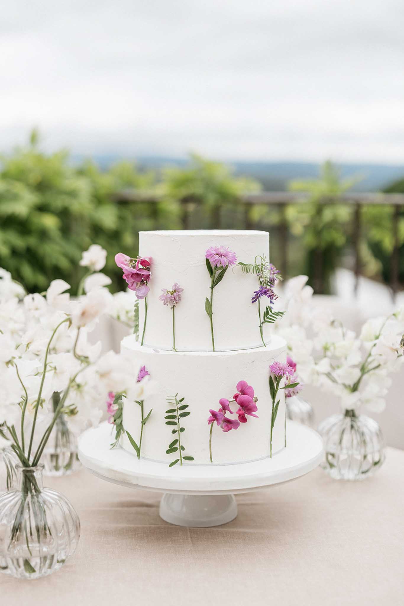 White wedding cake with purple flowers on pedestal stand at outdoor garden reception