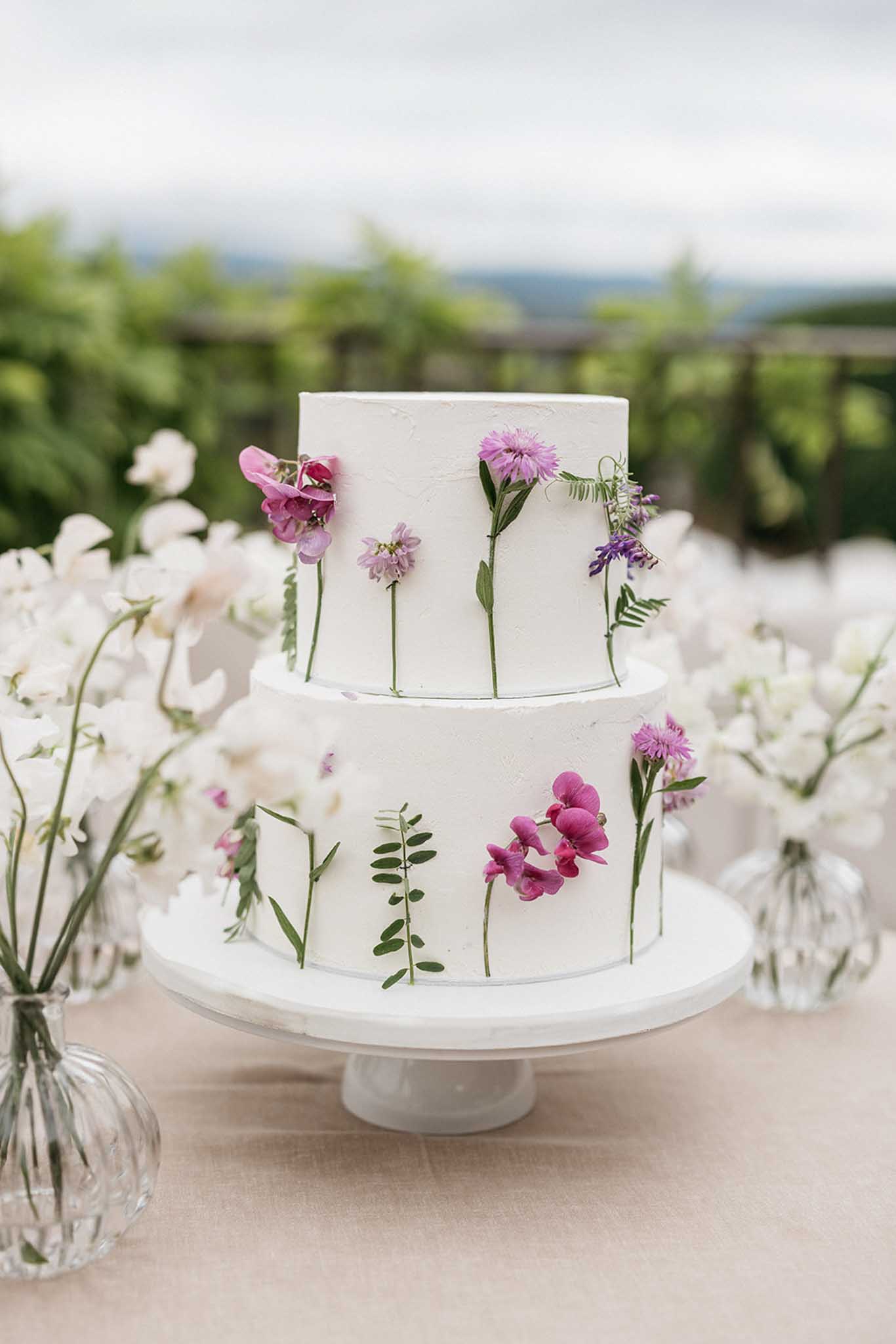 Two-tier wedding cake with purple flowers and greenery at outdoor countryside reception