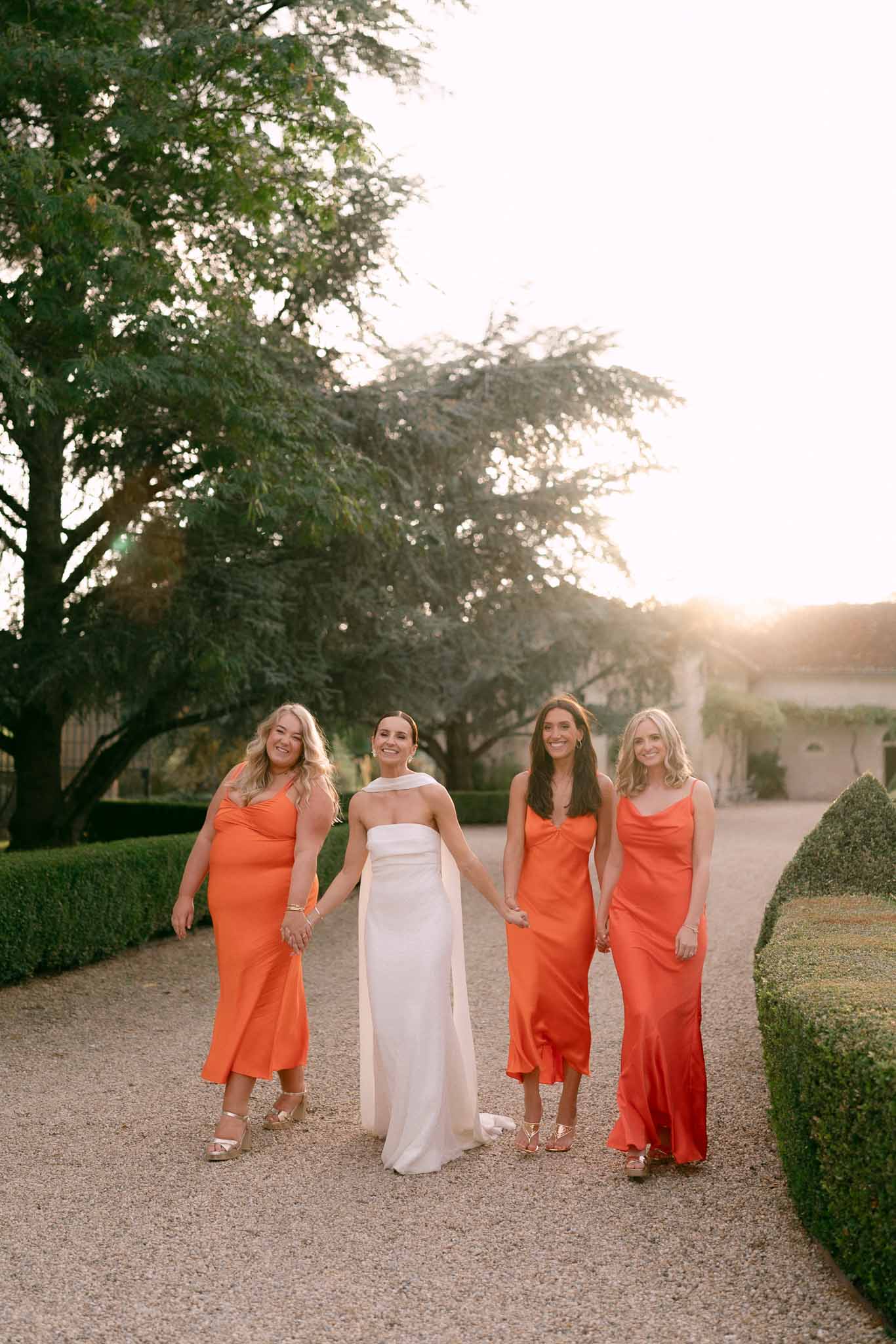 Bride in white column gown with three bridesmaids in orange and coral satin dresses at golden hour