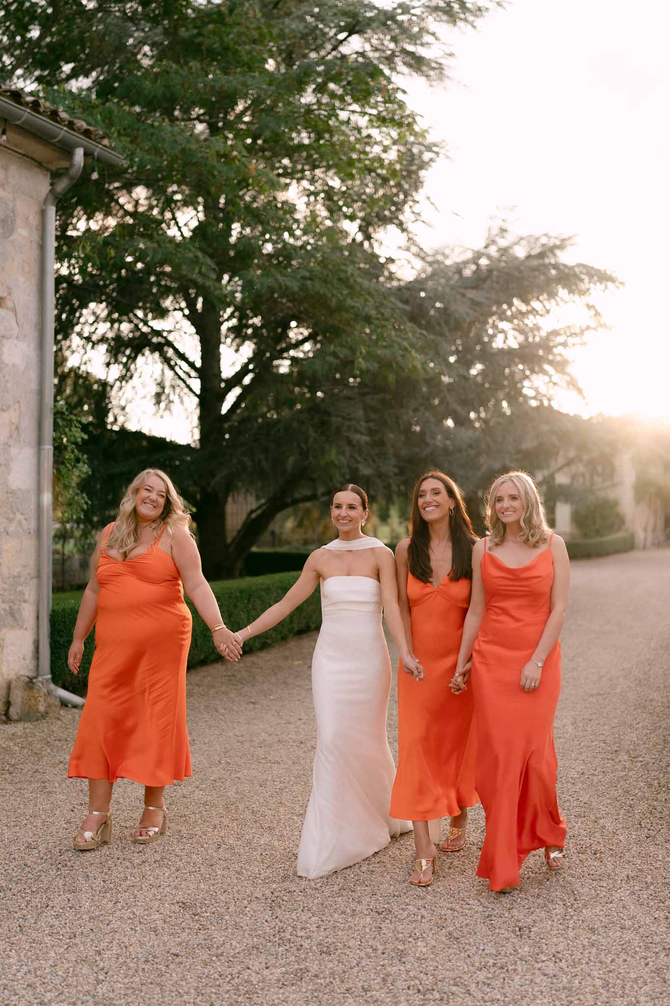 Bride in ivory halter gown walking with three bridesmaids in orange satin dresses at golden hour