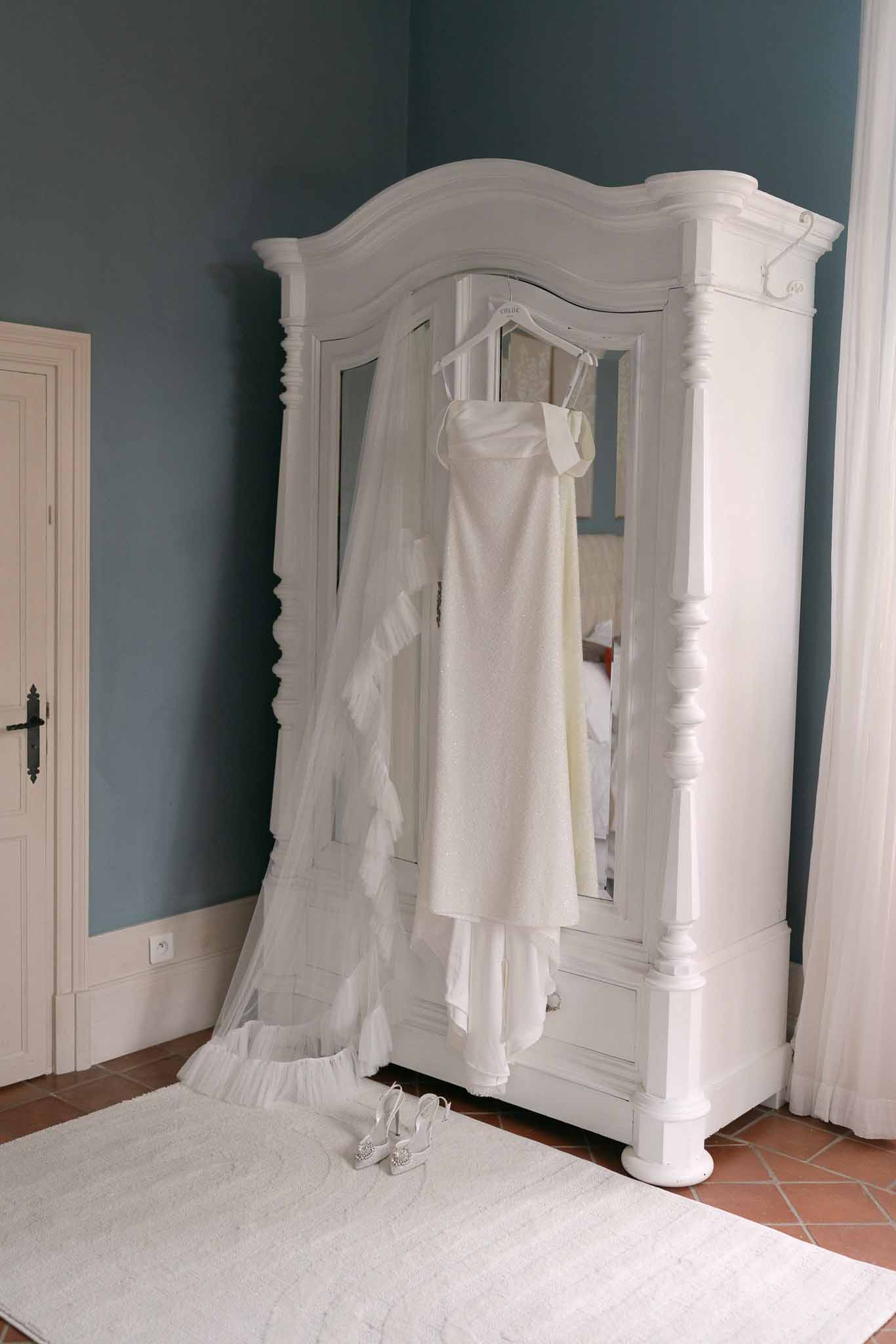 Wedding dress on personalized hanger with cathedral veil and bridal shoes on ornate French armoire