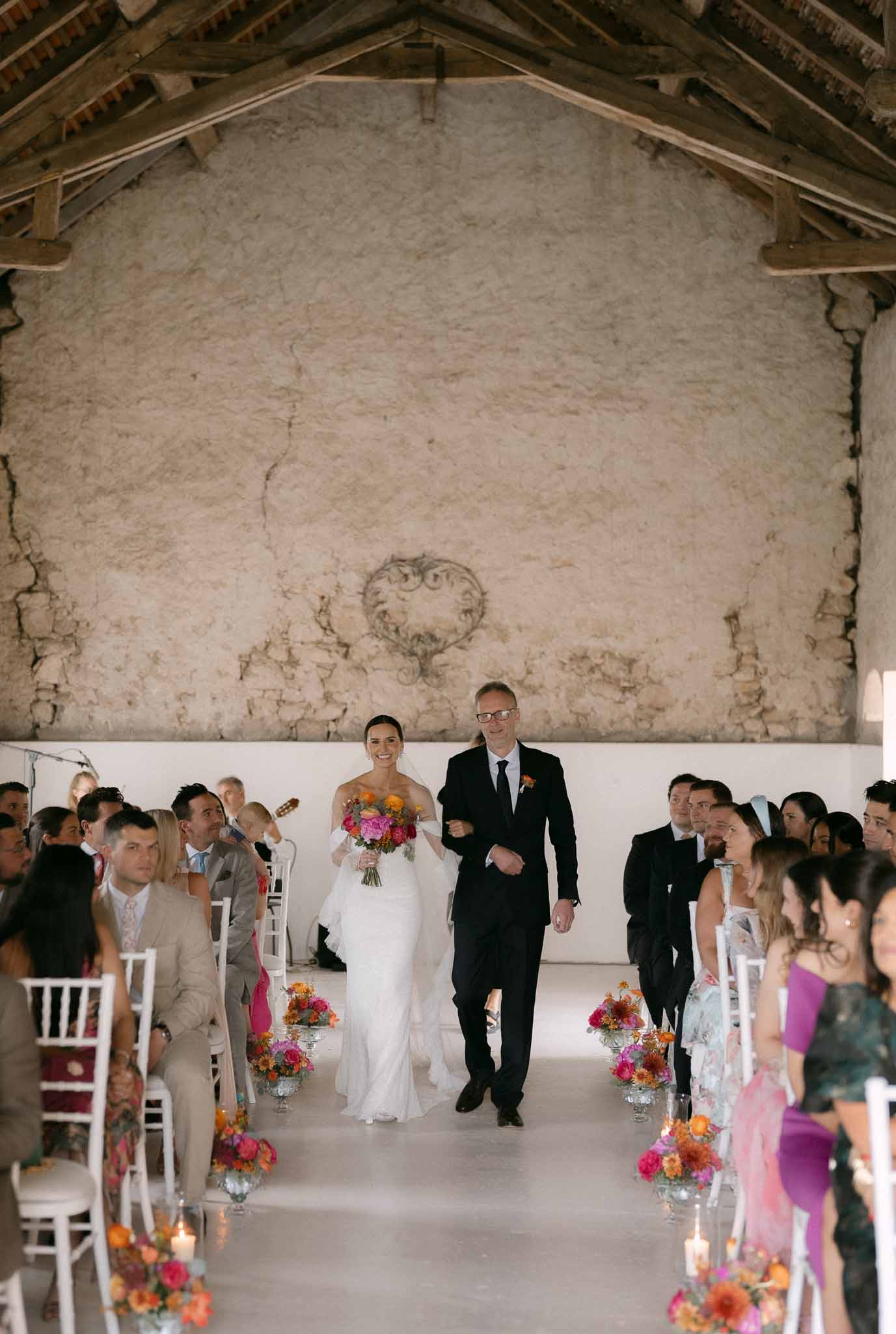 Bride walking down aisle with father in rustic barn venue with hot pink and orange floral arrangements and white chiavari ...
