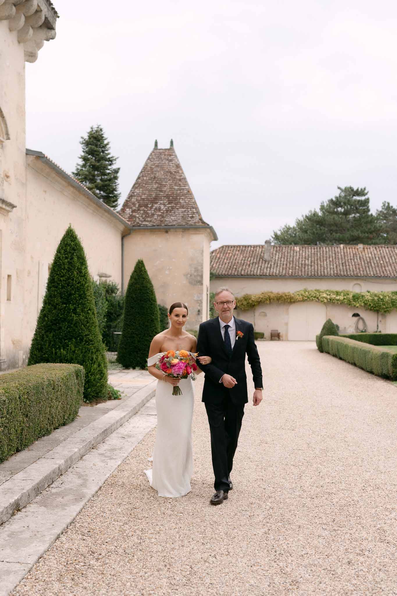 Bride in ivory column gown with hot pink and coral bouquet escorted by father through topiary-lined courtyard