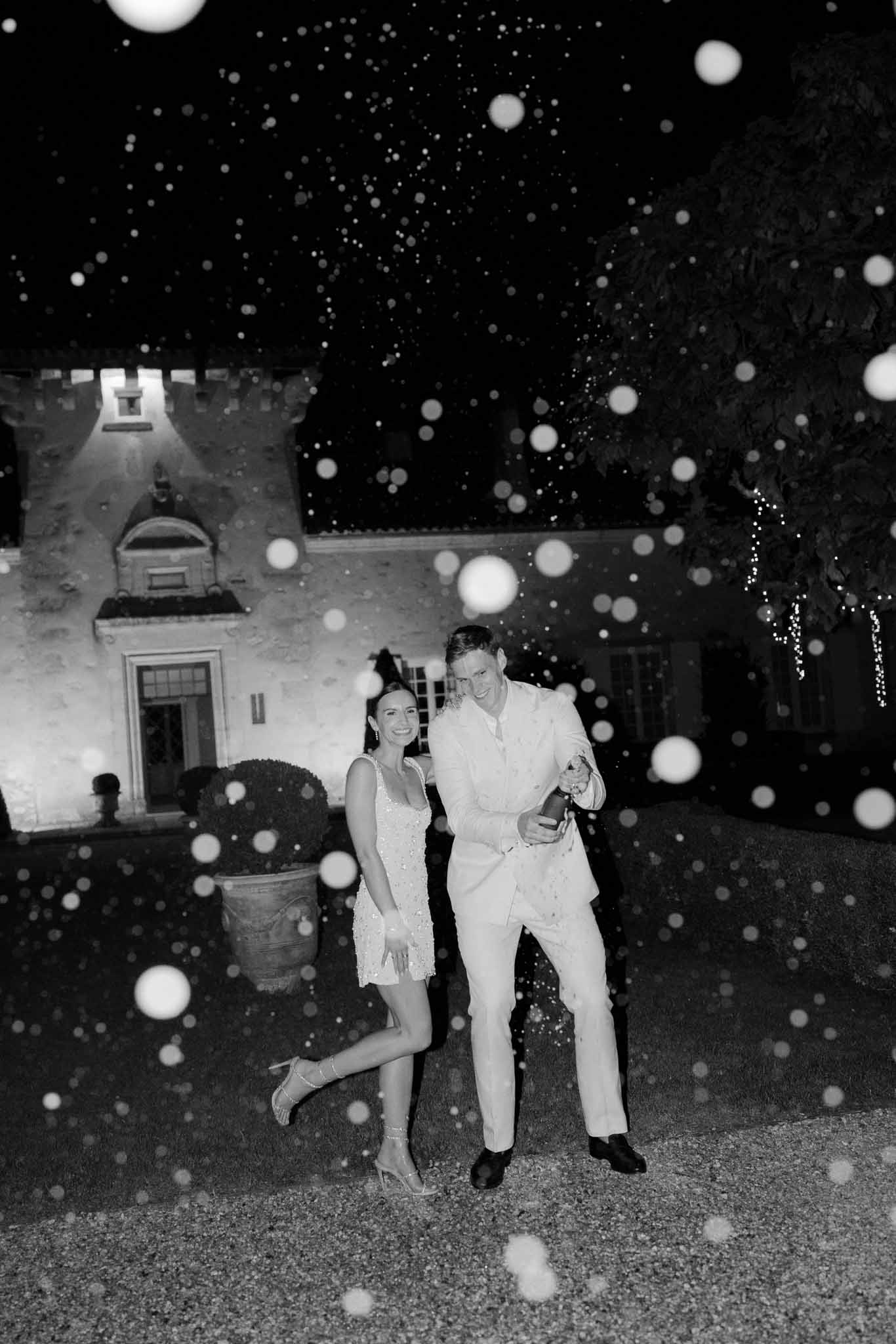 Black and white nighttime portrait of bride in mini dress and groom with champagne bottle laughing outside lit chateau