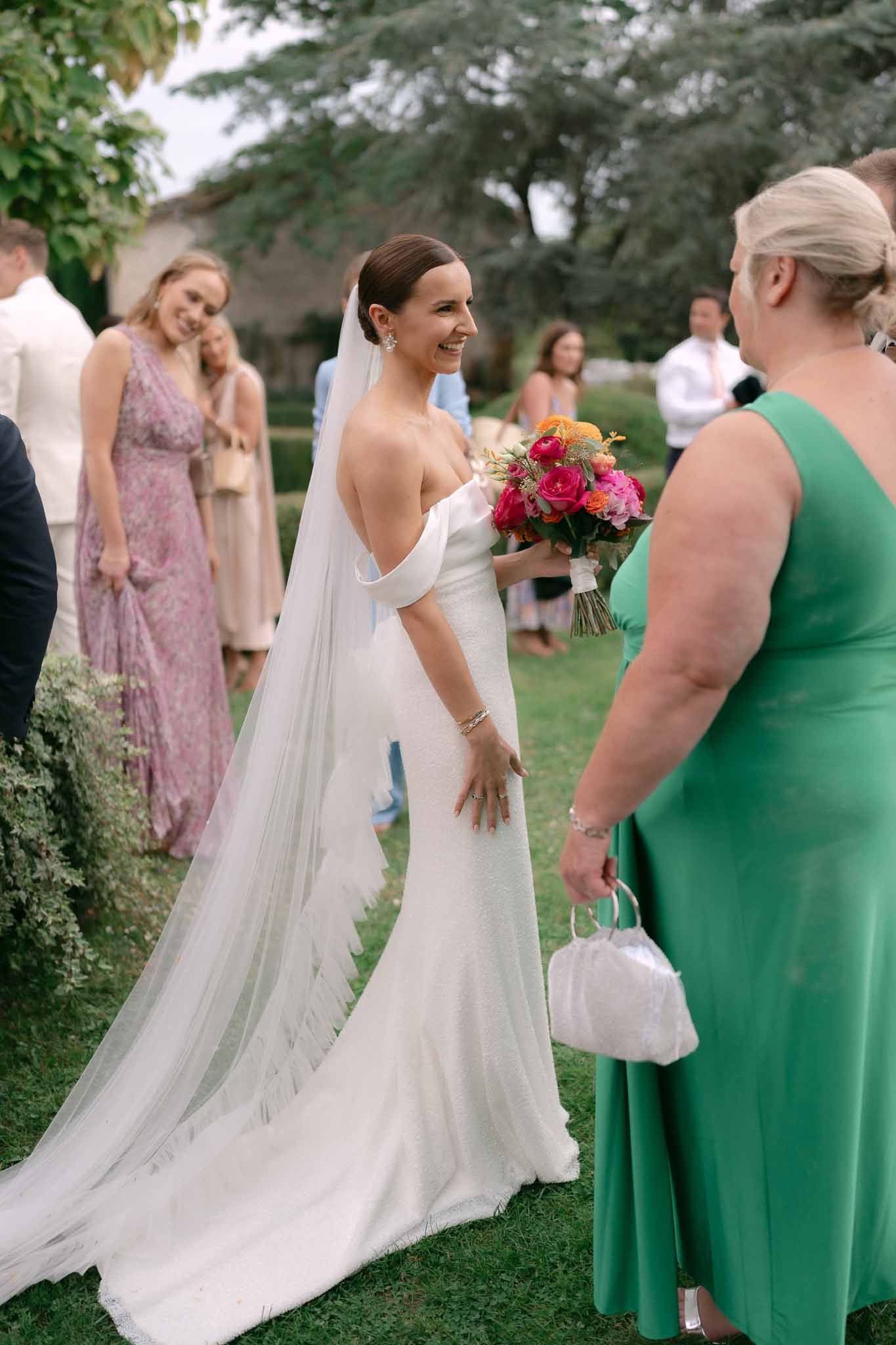 Bride in off-the-shoulder gown and cathedral veil holding pink and coral bouquet greeting guests during outdoor cocktail hour