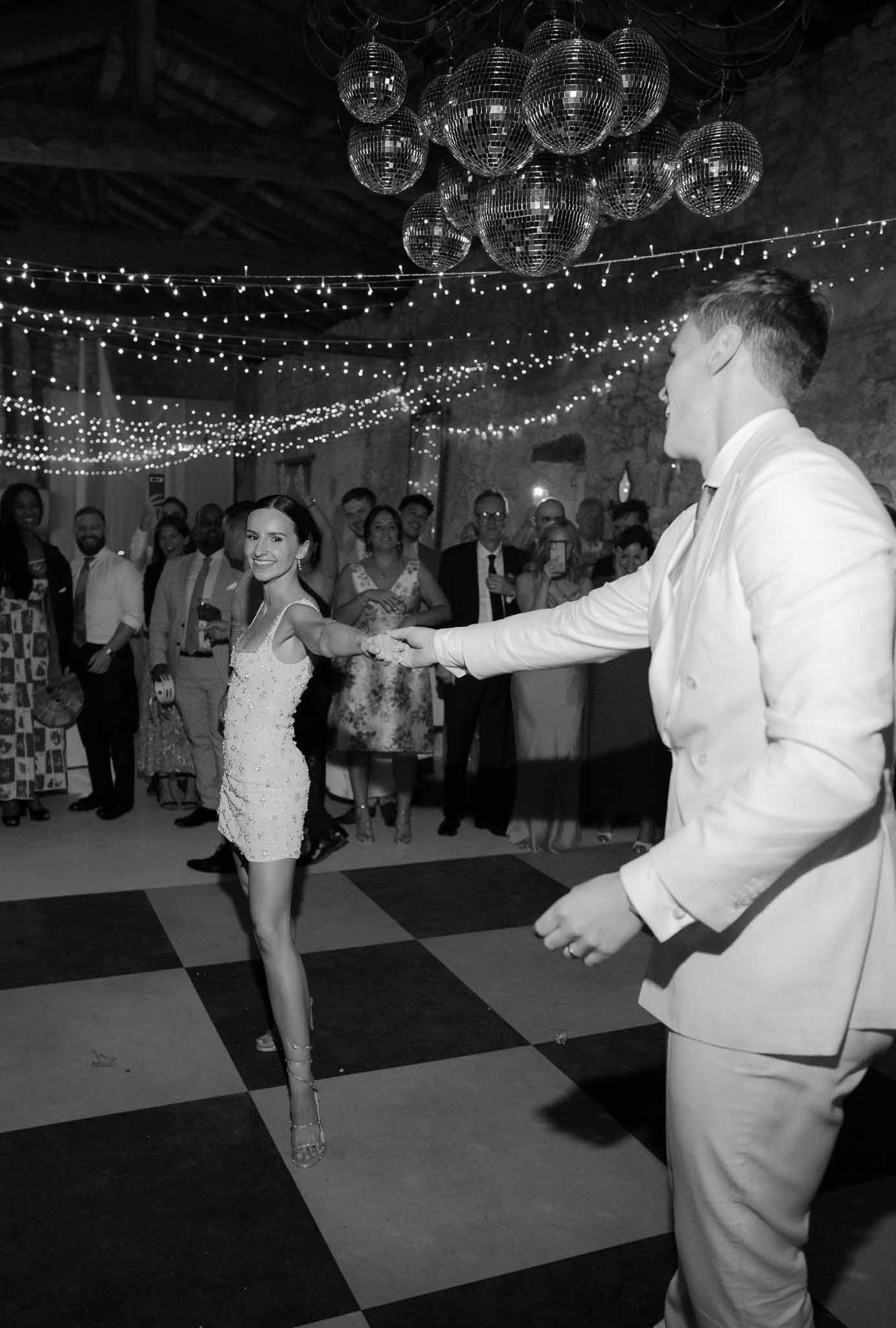 Black and white first dance on checkerboard floor under cluster of disco balls and fairy lights