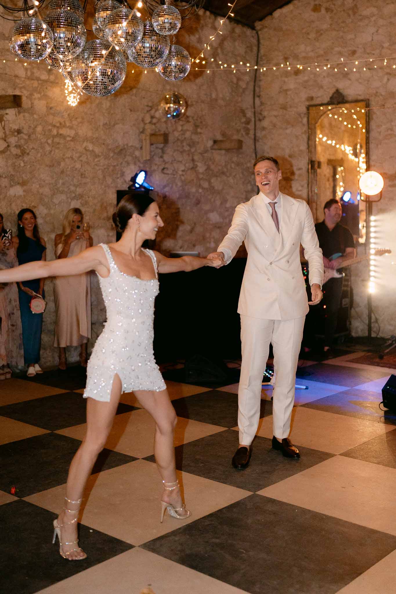 Bride in sequined mini dress and groom dancing on checkered floor under disco balls and fairy lights