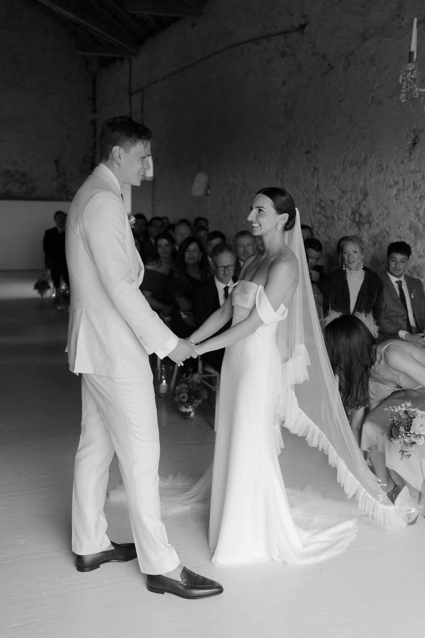 Black and white of couple holding hands at altar in rustic stone barn with exposed beams and seated guests