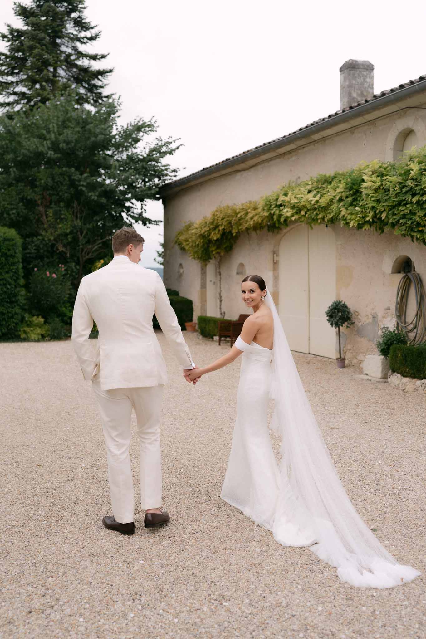 Bride looking back at camera with cathedral veil trailing on gravel as groom in cream suit walks ahead at stone property